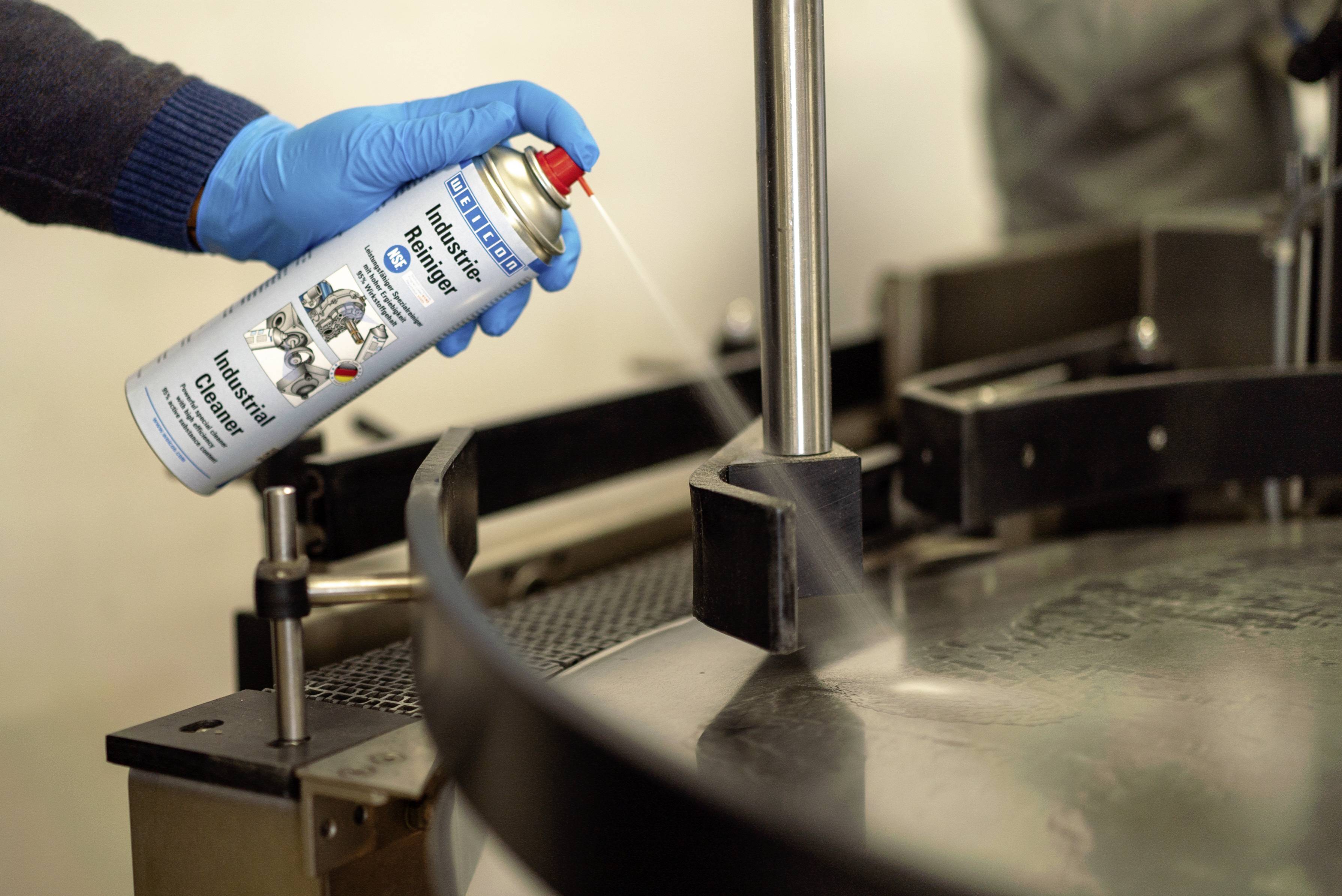 Eine Person mit blauen Handschuhen sprüht ein Schmiermittelspray auf ein Metallgerät in industrieller Umgebung.