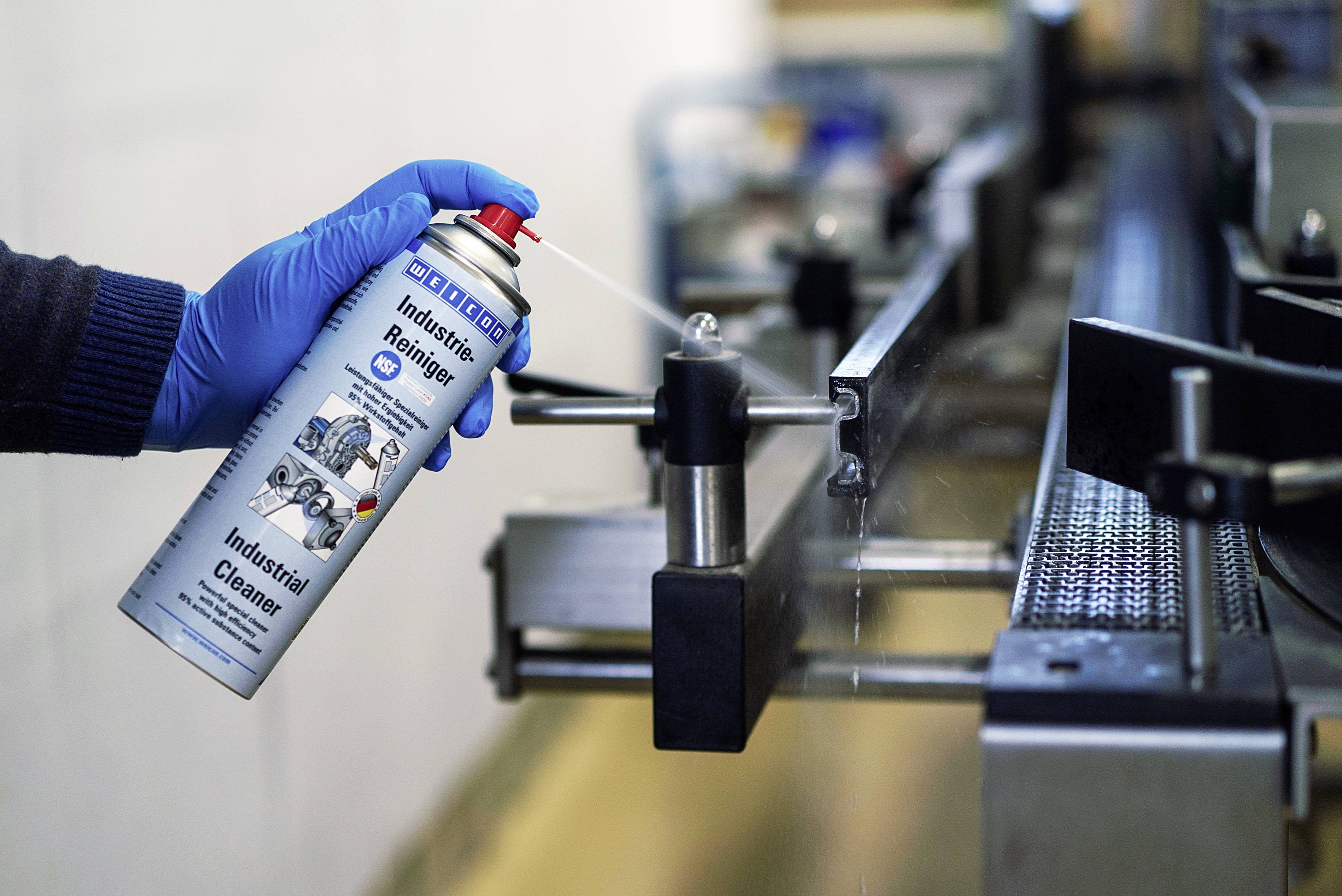 'Eine Person mit blauen Handschuhen sprüht Industriereiniger aus einer Dose auf eine Metallmaschine zur Wartung oder Reinigung.'
