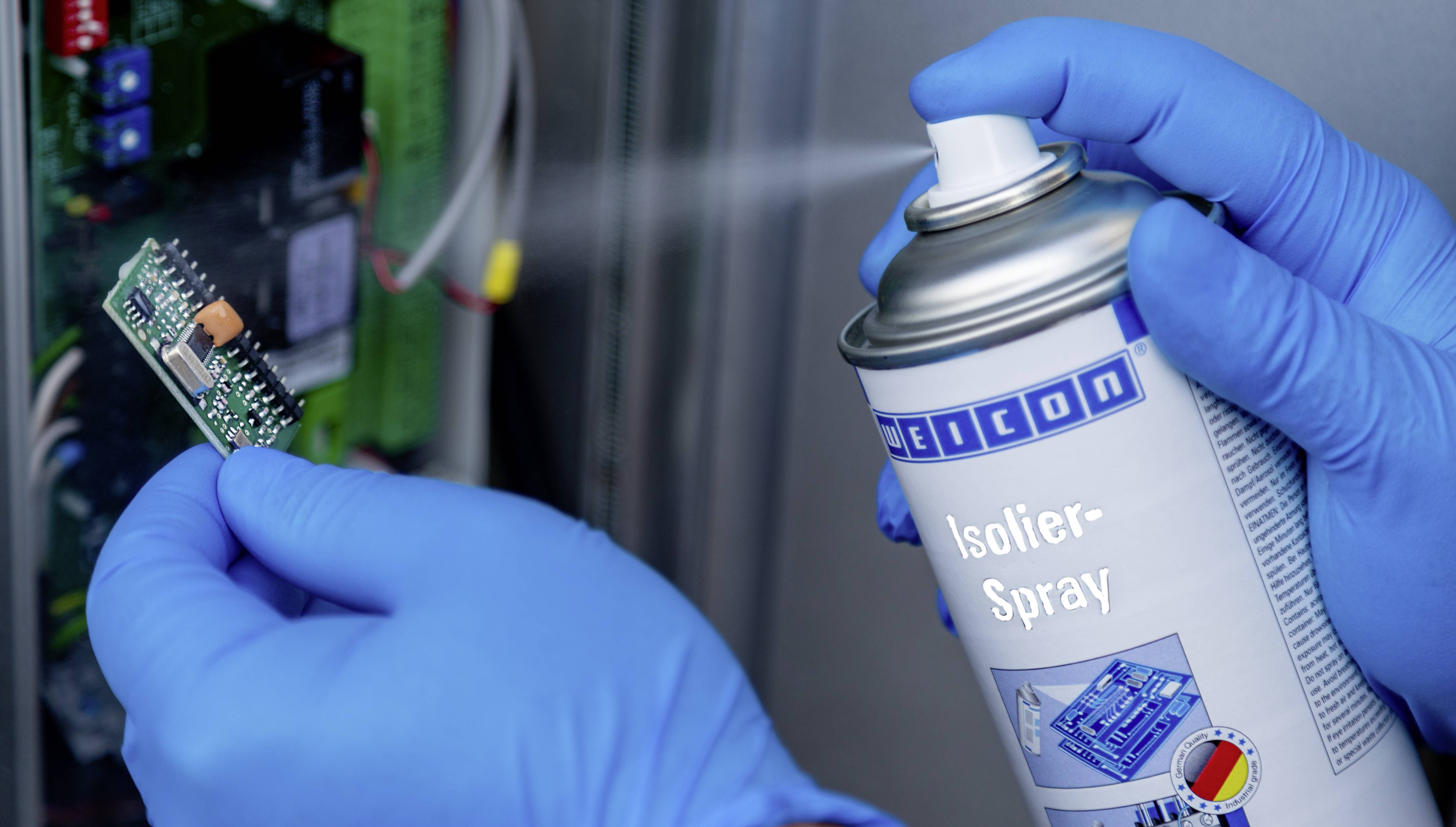 Eine Person mit blauen Handschuhen sprüht Isolier-Spray auf eine kleine elektronische Platine.