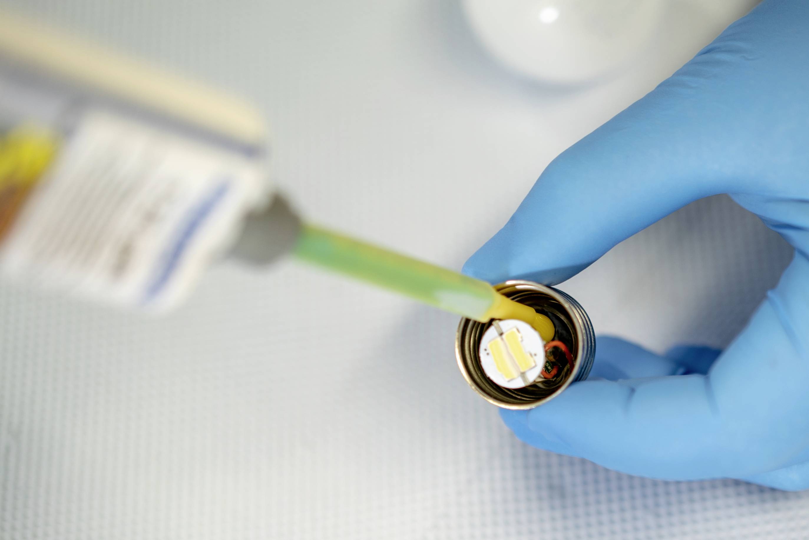 'Eine behandschuhte Hand hält einen kleinen Deckel, während Klebstoff aus einer Tube darauf aufgetragen wird.'