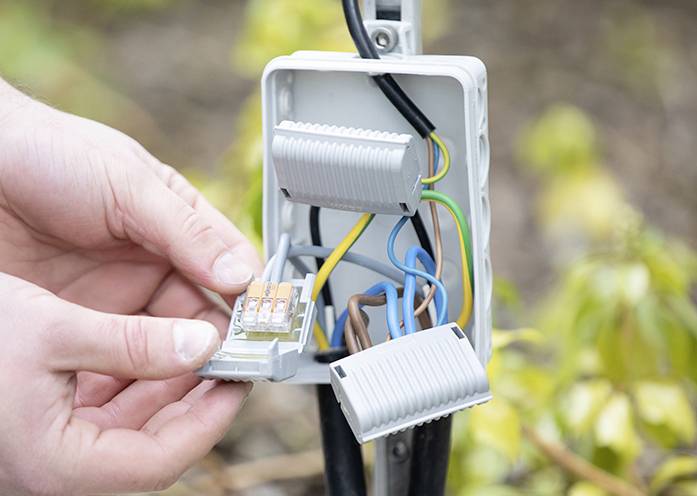 Person montiert elektrische Klemmen in einer grauen Anschlussdose im Freien. Bunte Drähte sind verbunden, Umgebung ist unscharf.