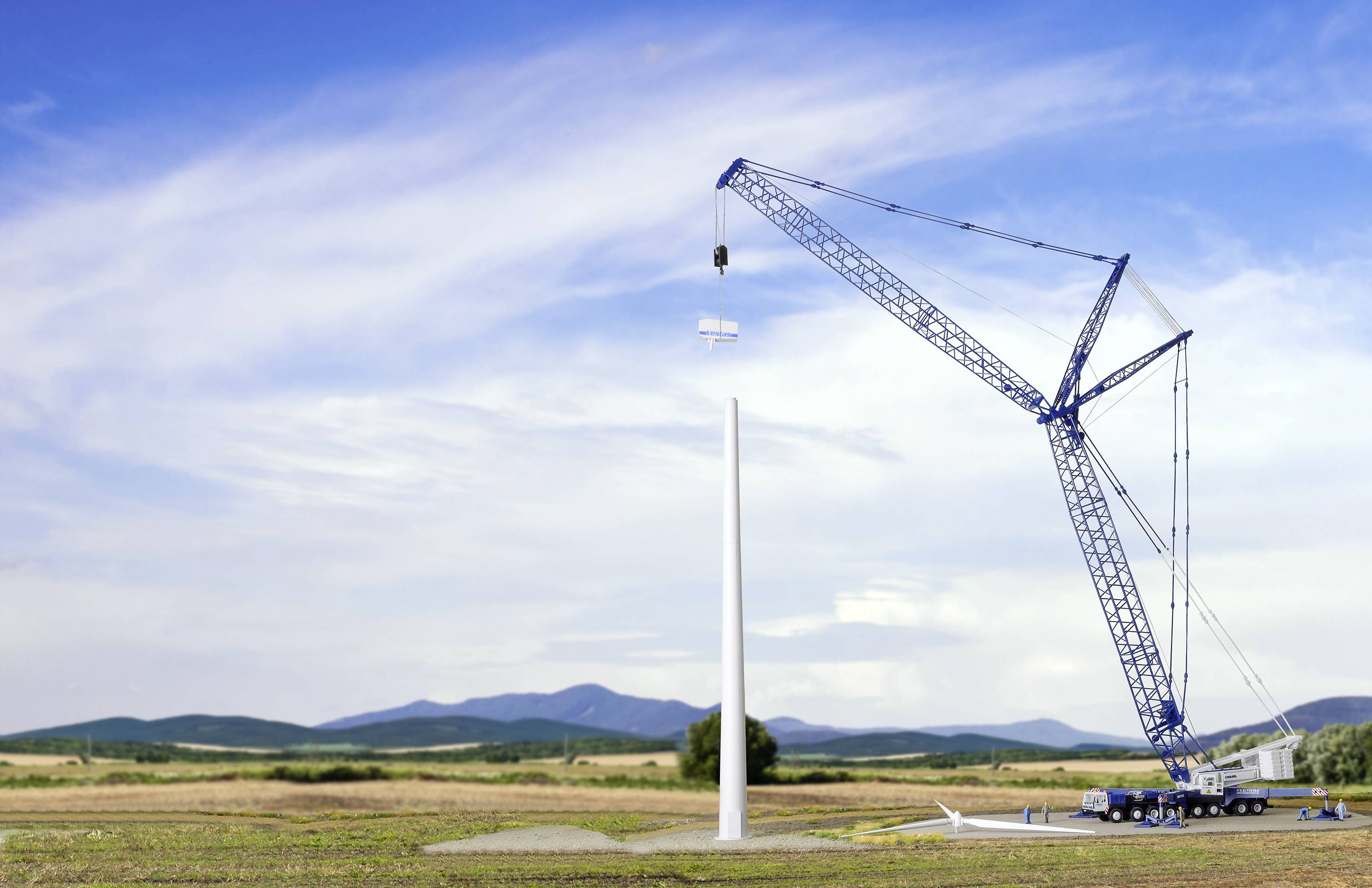 Ein großer blauer Kran errichtet eine Windturbine in einer weiten Landschaft. Der Kran hebt ein Bauteil an, während Berge im Hintergrund sichtbar sind.
