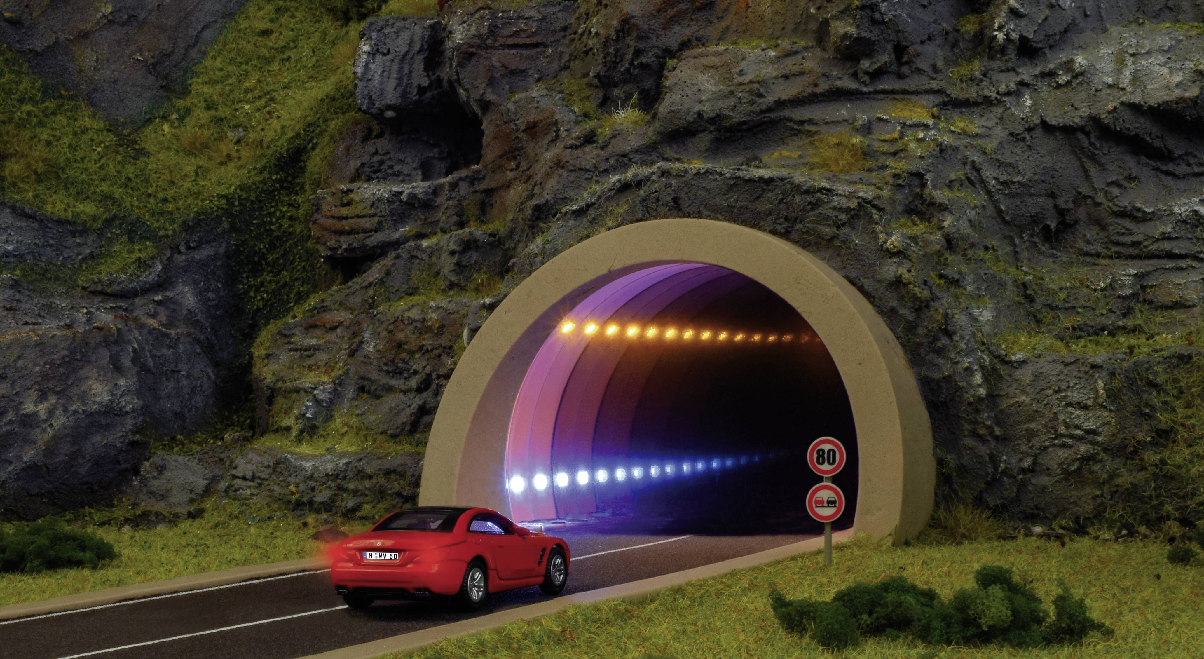 Ein rotes Auto fährt in einen beleuchteten Tunnel in hügeliger Landschaft. Verkehrszeichen zeigen '80 km/h' als Geschwindigkeitsbegrenzung an.