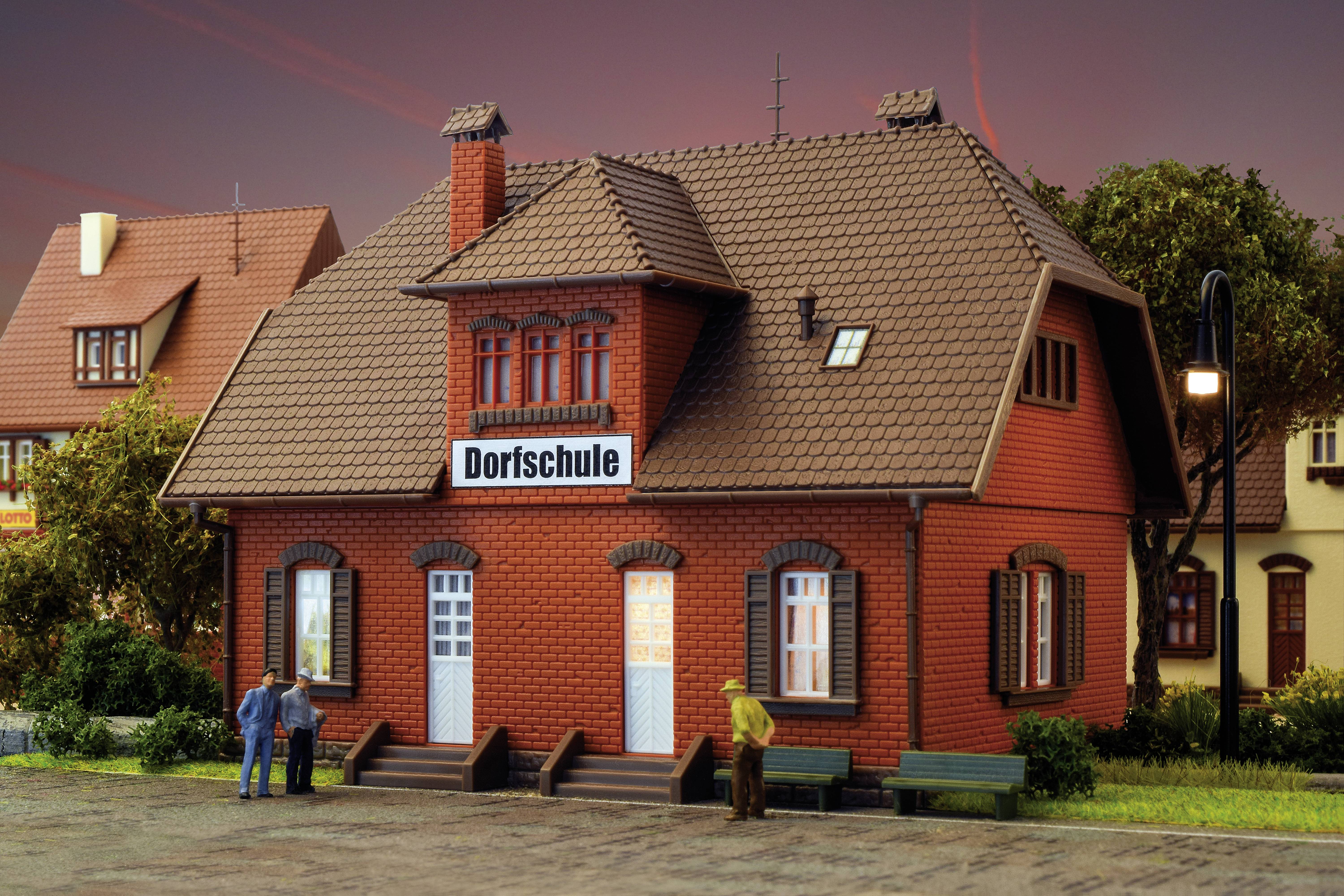 Ein kleines rotes Gebäude mit der Aufschrift 'Dorfschule' über dem Eingang, umgeben von Bäumen; Menschen stehen davor.