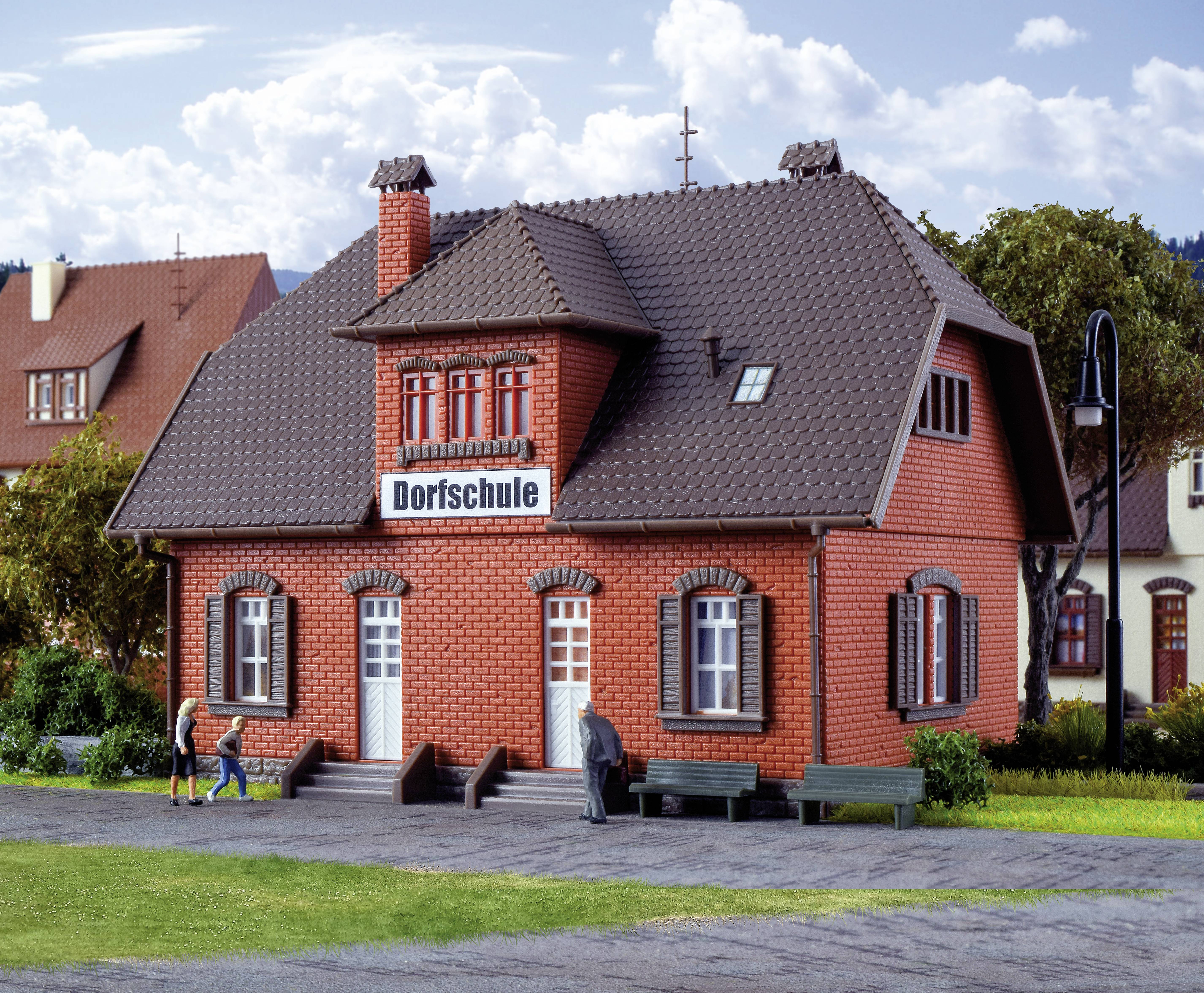 Rotes Backsteingebäude mit einem Schild 'Dorfschule' über den Türen, zwei Personen und eine Bank im Vordergrund.