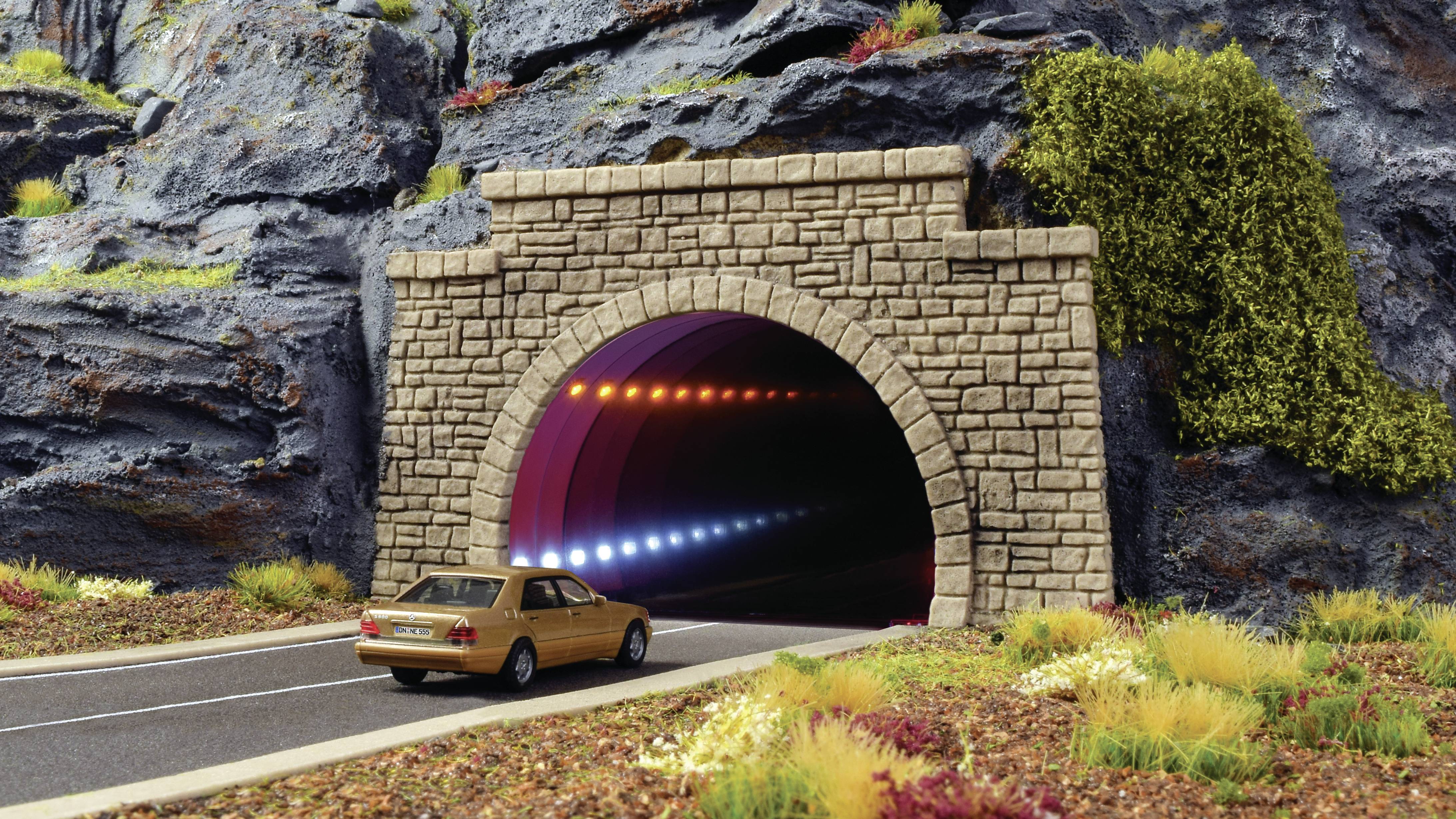 Ein gelbes Auto fährt in einen Steintunnel, umgeben von einer Landschaft mit buntem Laub. Im Tunnel sind bunte Leuchten sichtbar.