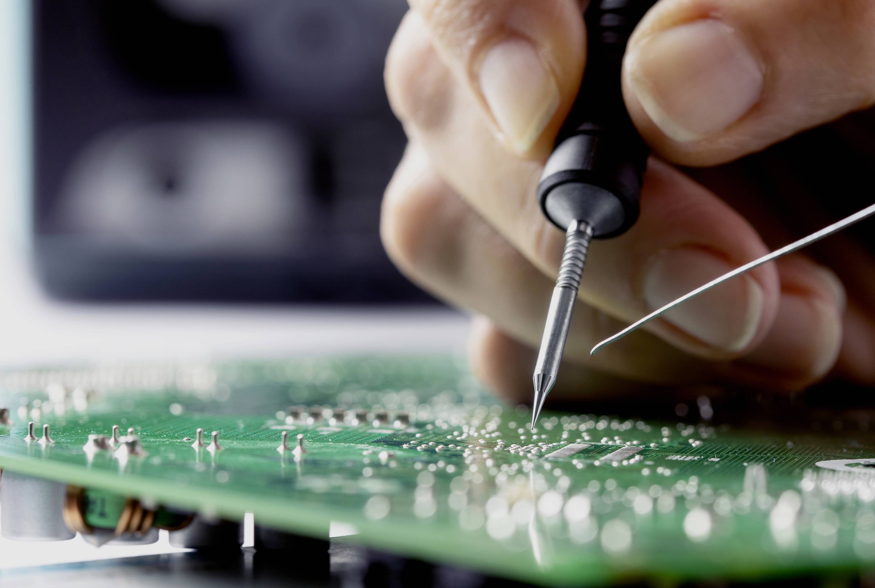Eine Hand lötet mit einem feinen Lötkolben eine Leiterplatte. Nahaufnahme zeigt präzise Elektronikarbeit auf grünem Board.