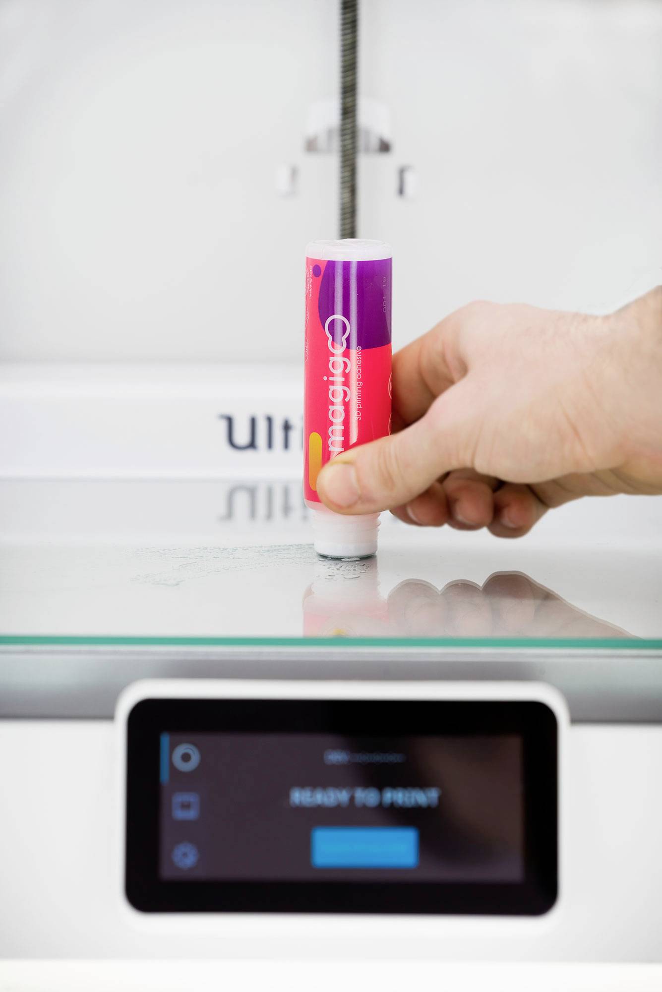 Eine Hand hält eine Leimstift-Verpackung vor einem 3D-Drucker. Der Bildschirm des Druckers zeigt 'Ready to Print'.