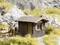 Eine kleine Holzhütte mit Schild 'Berghütte H. Nuottokari', umgeben von felsiger Landschaft. Zwei Personen mit Rucksäcken und Hund in der Nähe.