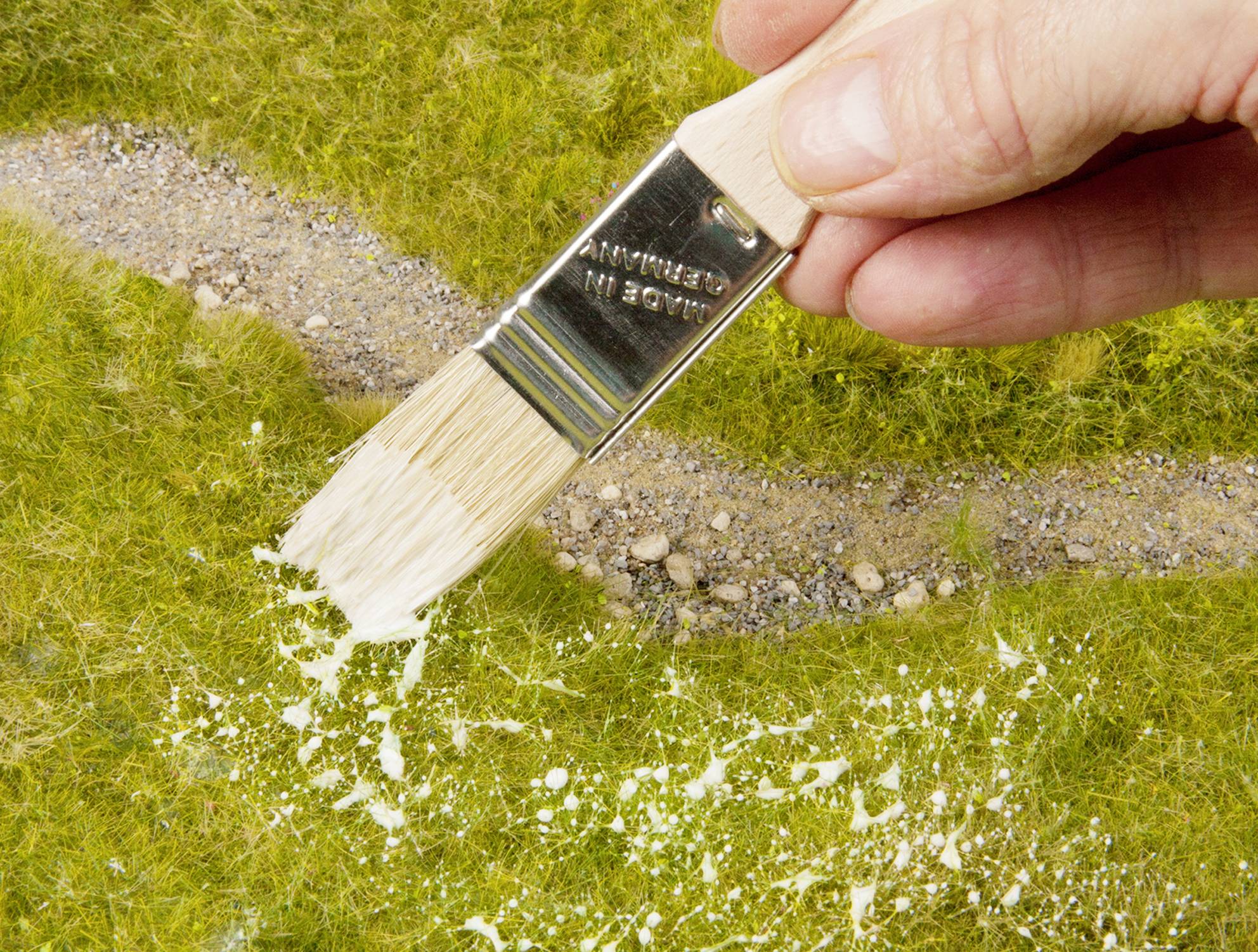 Eine Hand hält einen Pinsel und streicht weißes Material auf kleine Gras- und Kiesflächen, möglicherweise ein Modellbauprojekt.