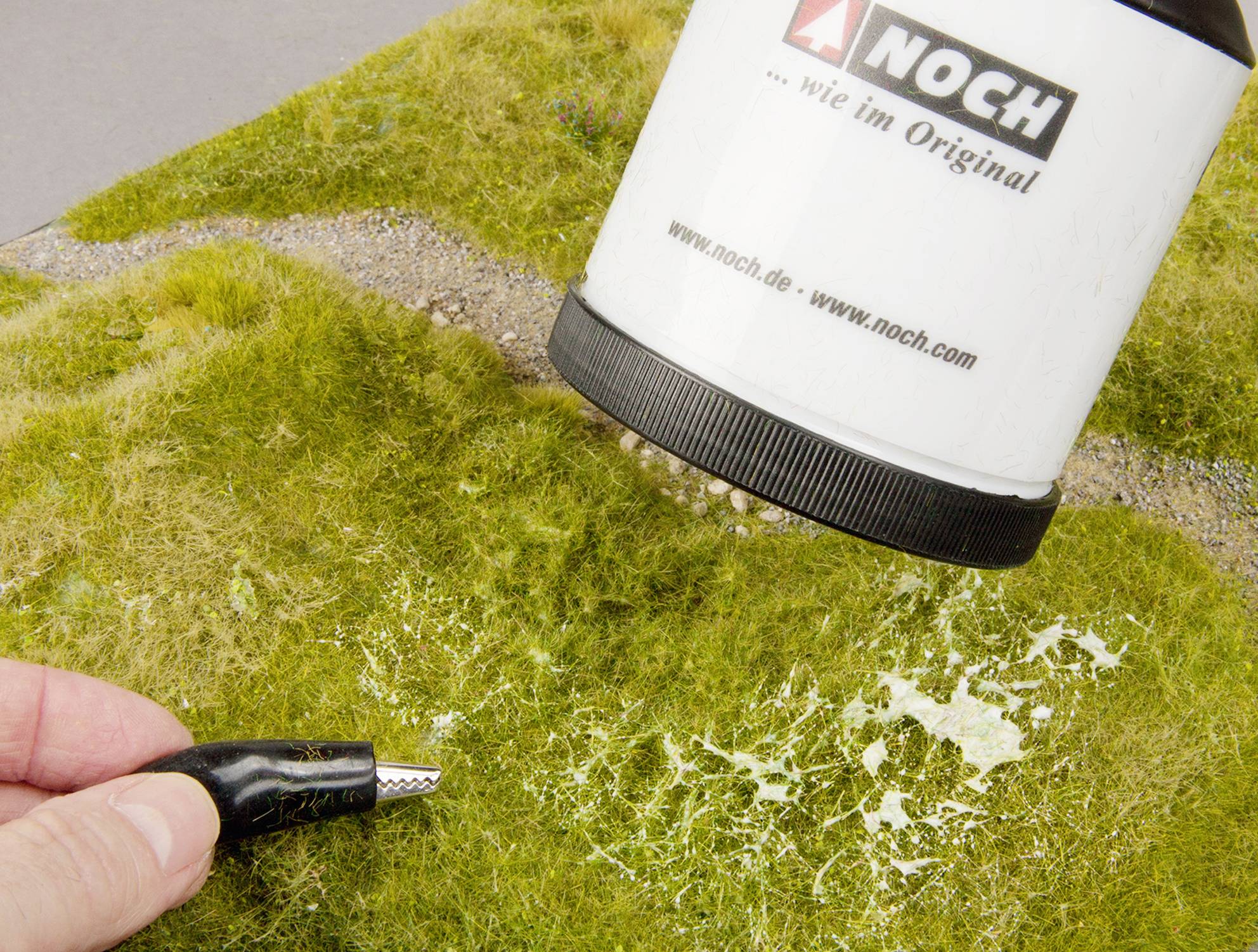 Ein Mensch hält ein Elektrokabel neben eine Flasche mit Sprühsystem, um Gras auf einer Modelllandschaft zu gestalten.