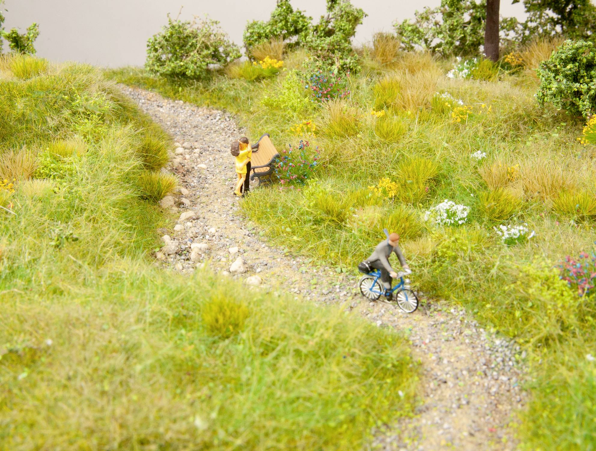 Ein Miniaturmodell zeigt einen Radfahrer auf einem Kiesweg durch eine grüne Landschaft mit Bäumen und Blumen, gefolgt von einem Wanderer mit einem Karton.