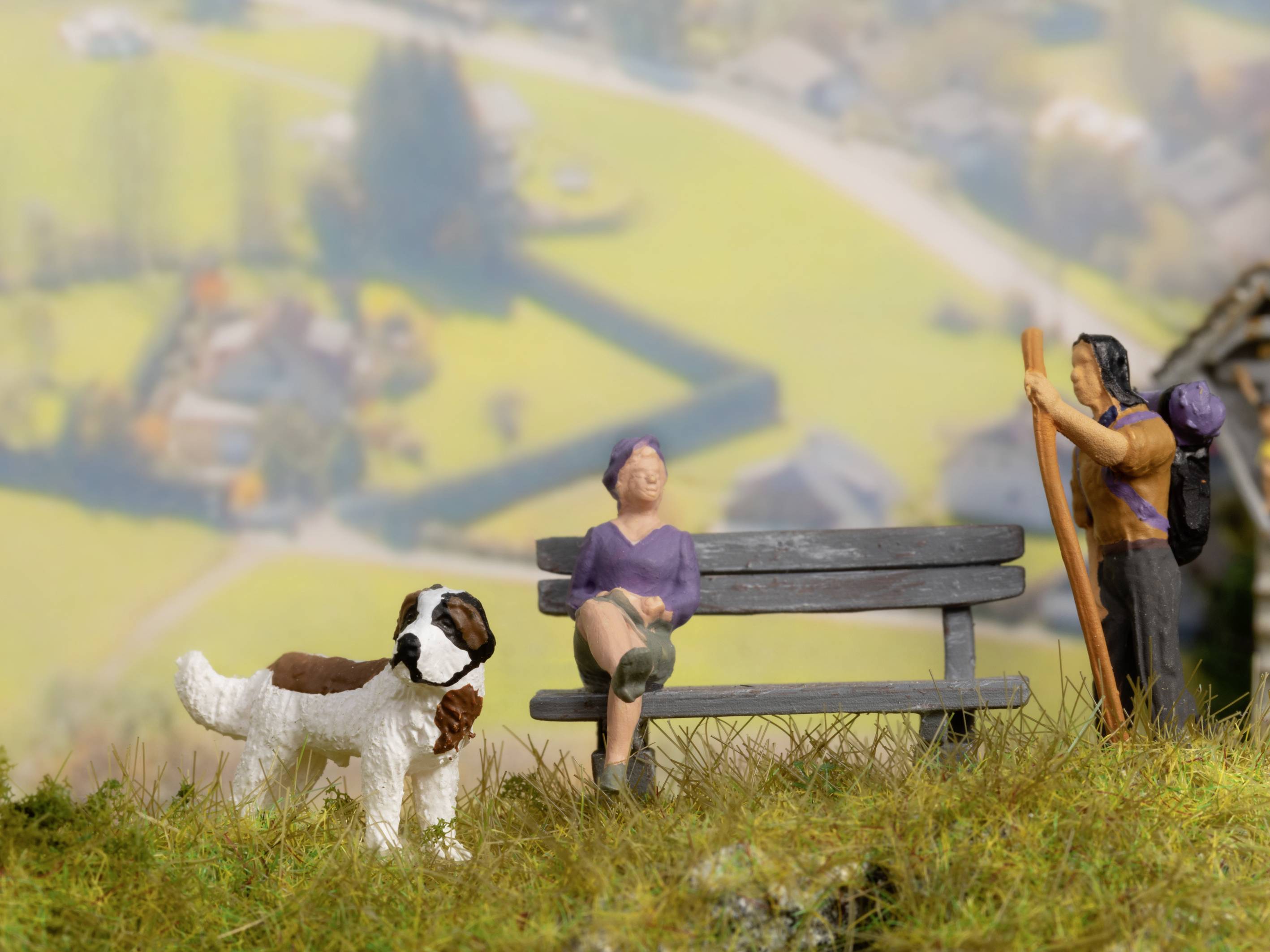 Zwei Figuren sitzen und stehen auf einer Bank auf grünem Rasen, mit einem Hund daneben. Hintergrund zeigt unscharfe Landschaft.
