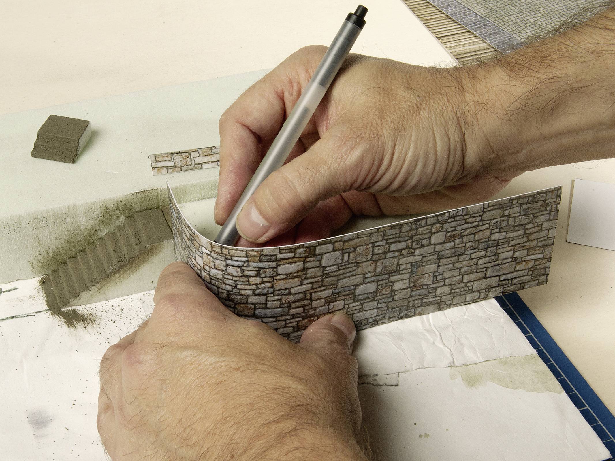 'Zwei Hände bauen ein Miniatur-Diorama, befestigen gedruckte Steinmauer an der Form mit einem Stift und Klebstoff.'