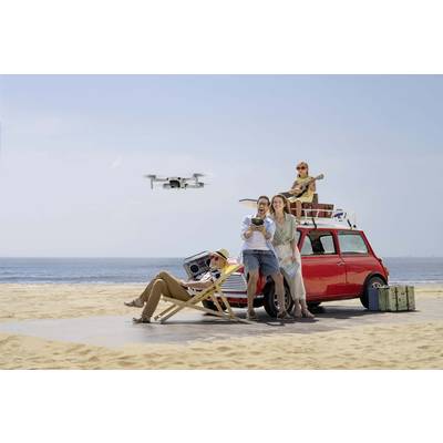 Eine Gruppe von vier Personen entspannt sich am Strand neben einem roten Auto. Ein Mann steuert eine Drohne, während andere entspannen.