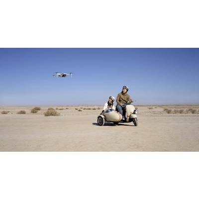 Zwei Personen sitzen auf einem Motorrad mit Beiwagen in einer Wüstenlandschaft; eine Drohne fliegt nahe bei ihnen am blauen Himmel.