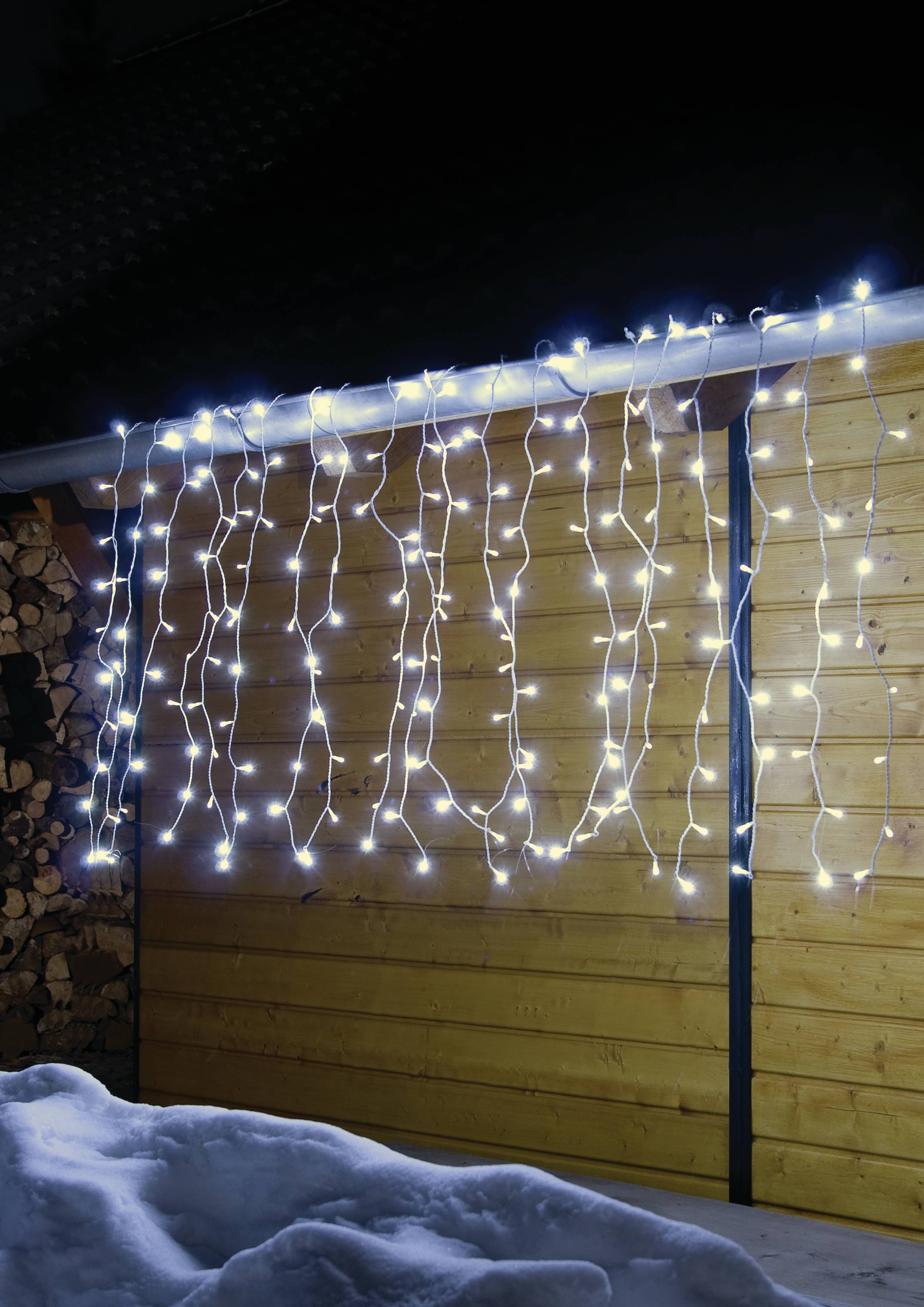 Lichterketten hängen an einem Holzgebäude bei Nacht, strahlen warmes Licht auf den umgebenden Schnee.