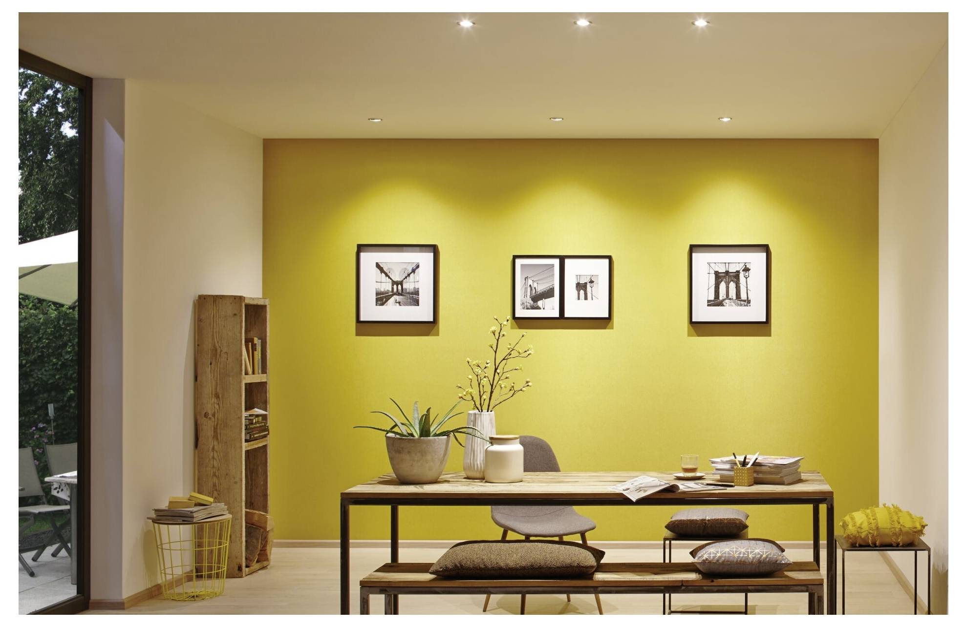 Wohnzimmer mit Holztisch, darauf Pflanzen und Bücher. Gelbe Akzentwand mit drei gerahmten Schwarz-Weiß-Fotos. Rechter Raumteil beleuchtet.