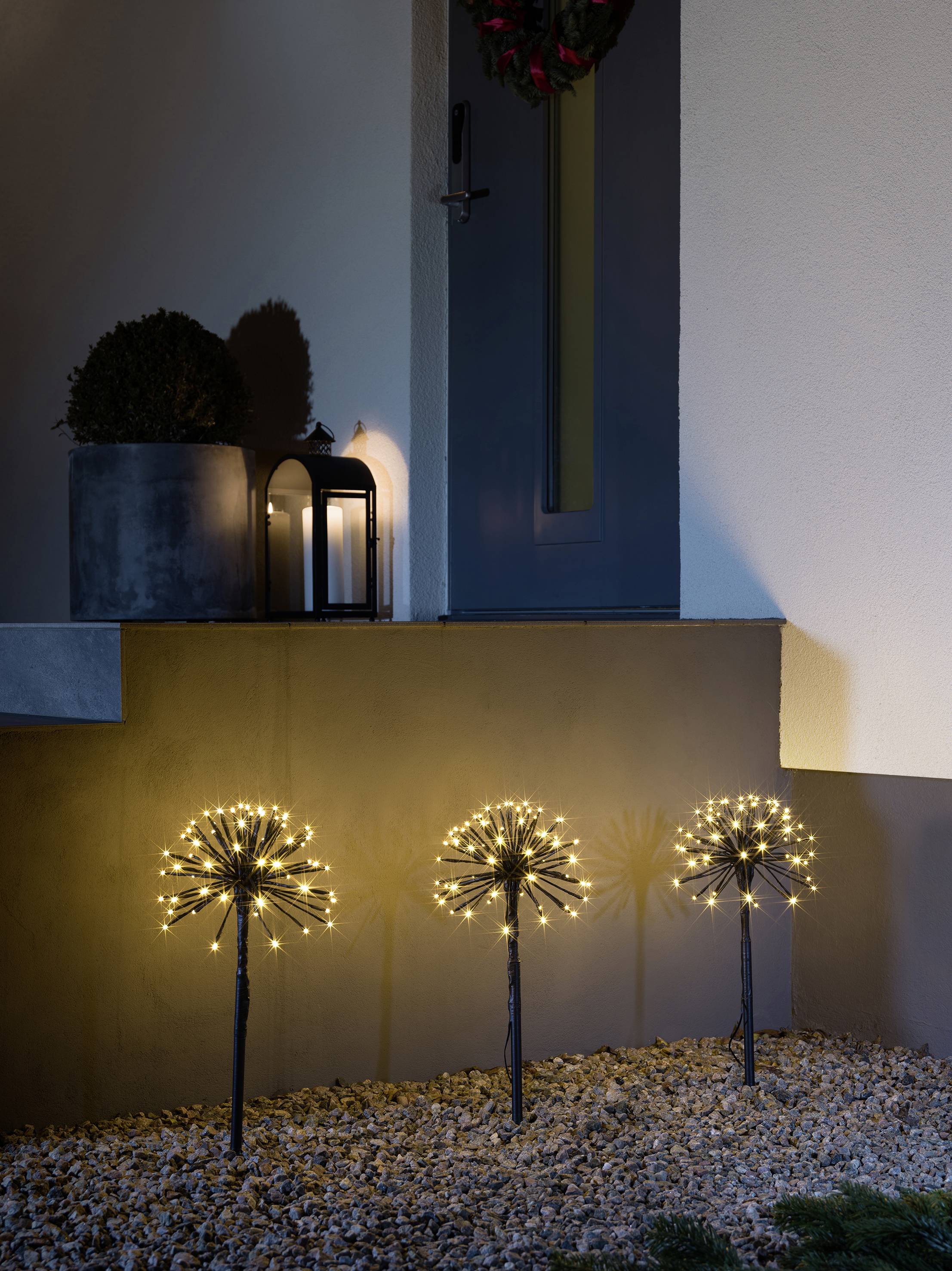 Im Vorgarten sind dekorative Lichter in Form von Blumen angeordnet, die bei Nacht leuchten. Im Hintergrund steht eine Laterne.