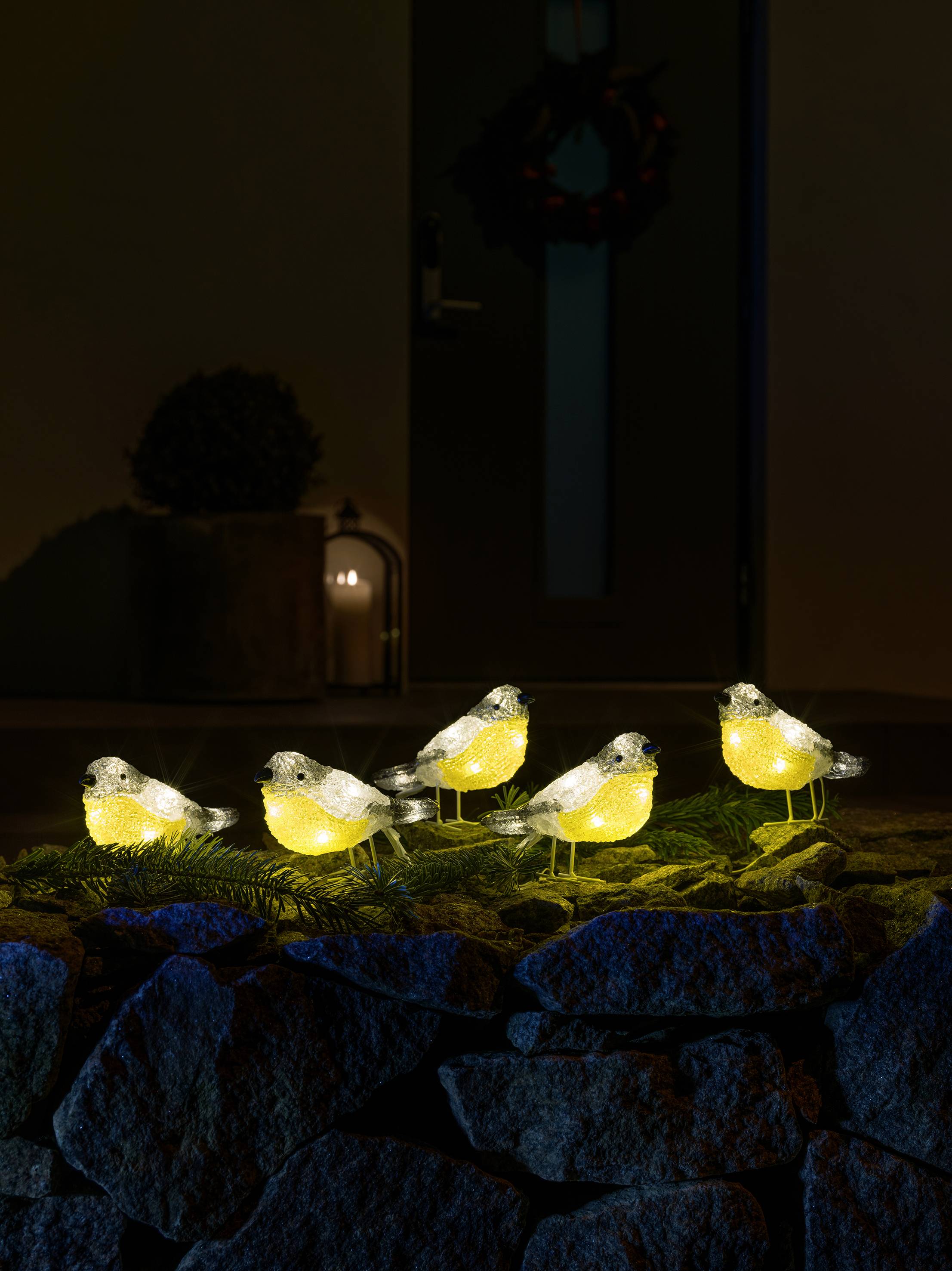 Dekorative Beleuchtung in Form von fünf gelben und weißen Vögeln auf einem Steingarten, vor einem Haus im Hintergrund.