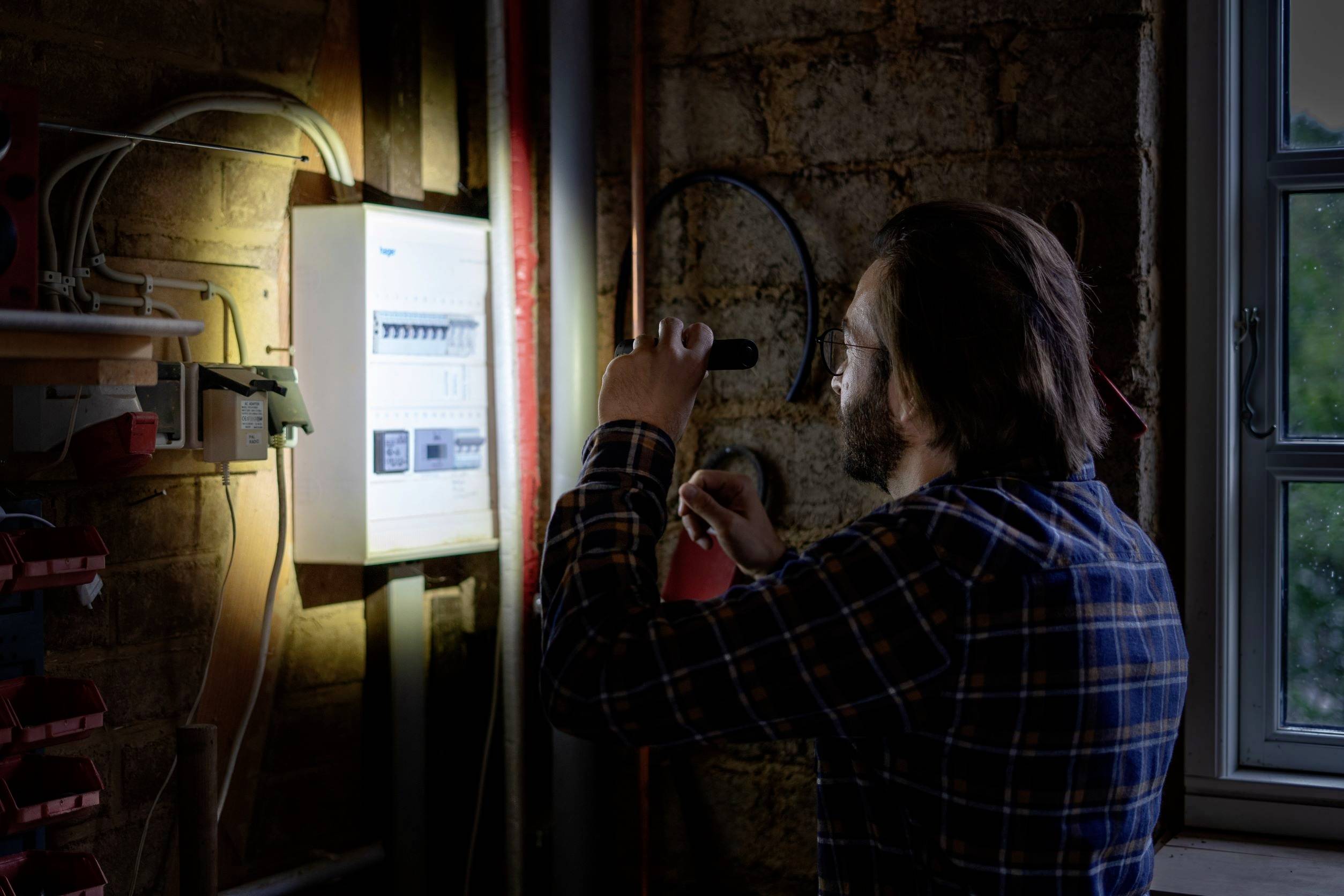 Ein Mann in kariertem Hemd untersucht bei schwachem Licht mit einer Taschenlampe einen Sicherungskasten an einer Ziegelwand.