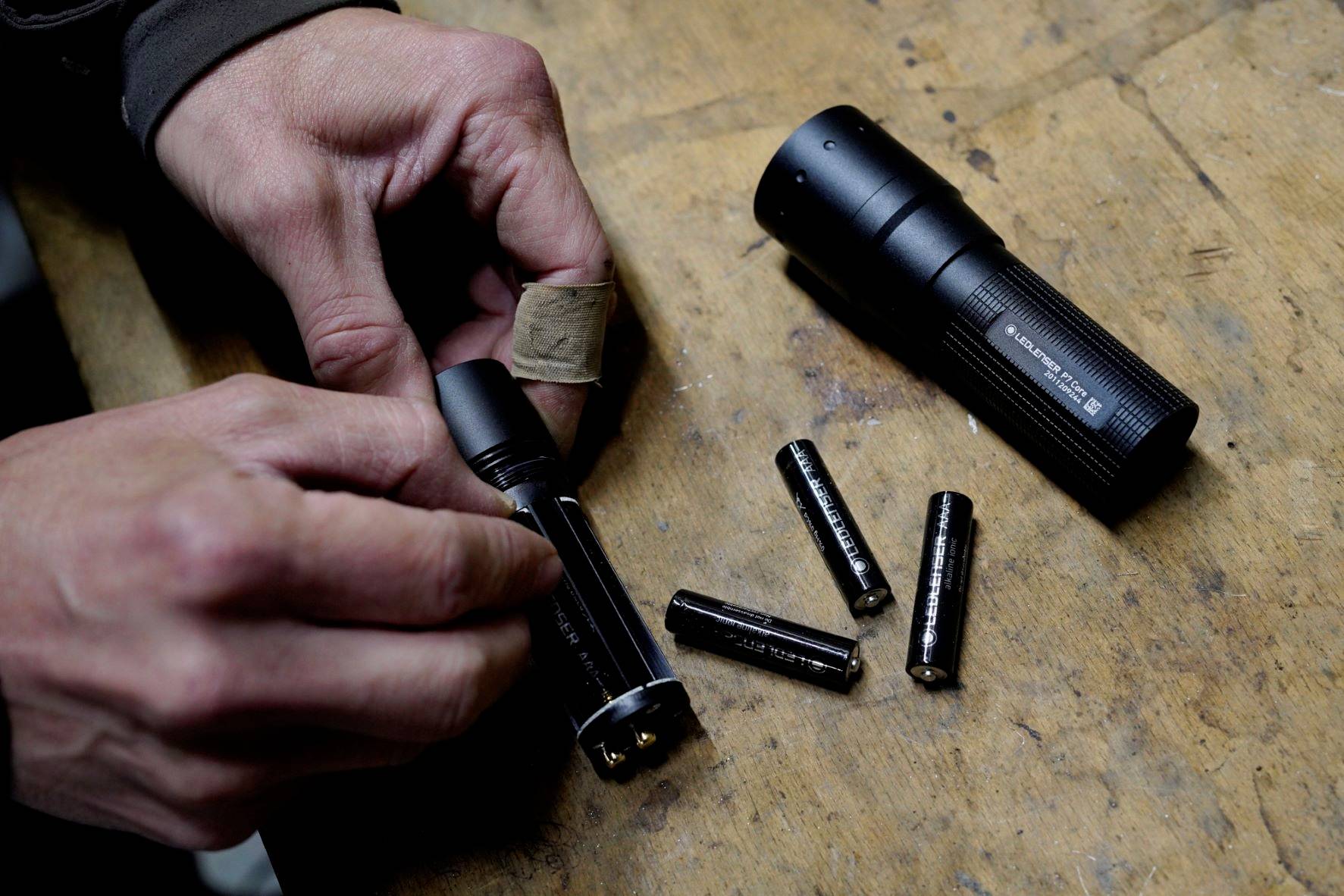 Eine Person wechselt Batterien in einer schwarzen Taschenlampe auf einem Holztisch. Vier AA-Batterien liegen daneben.