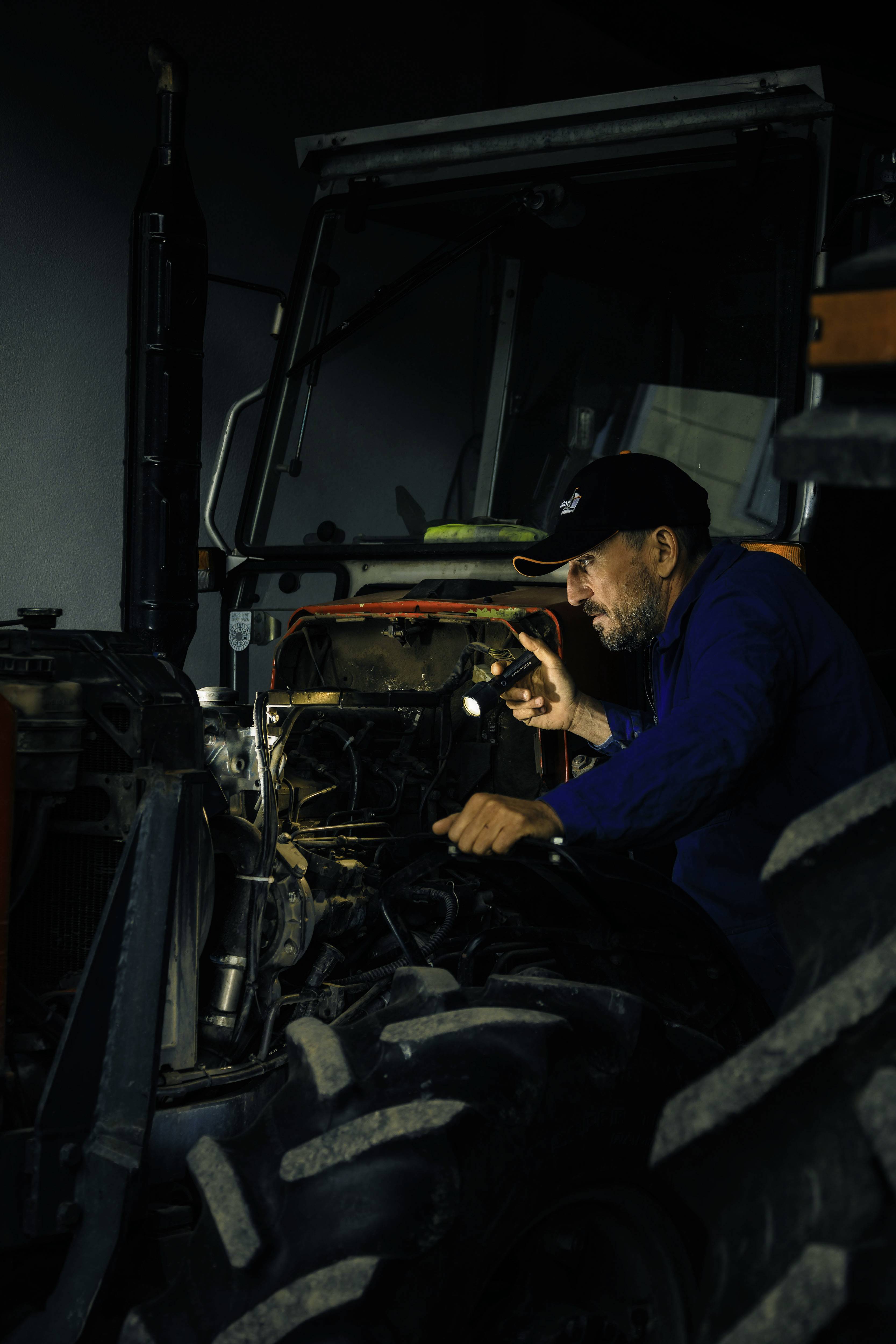 Ein Mann repariert einen Traktor in einer dunklen Werkstatt, beleuchtet von einer Taschenlampe. Er konzentriert sich auf den Motor.