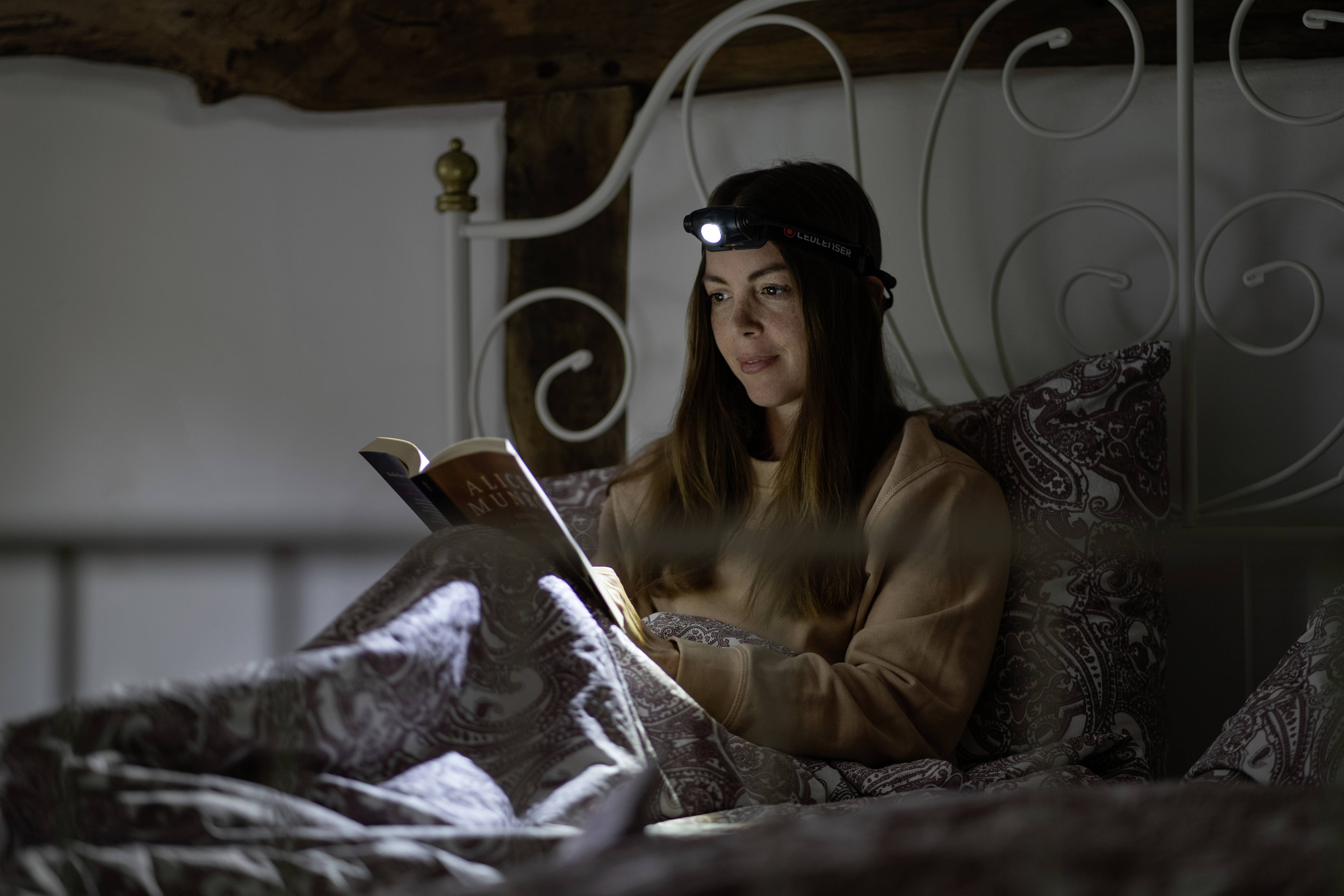 Frau liest im Bett ein Buch, trägt eine Stirnlampe. Schlafzimmer mit dekorativen Bettpfosten im Hintergrund.