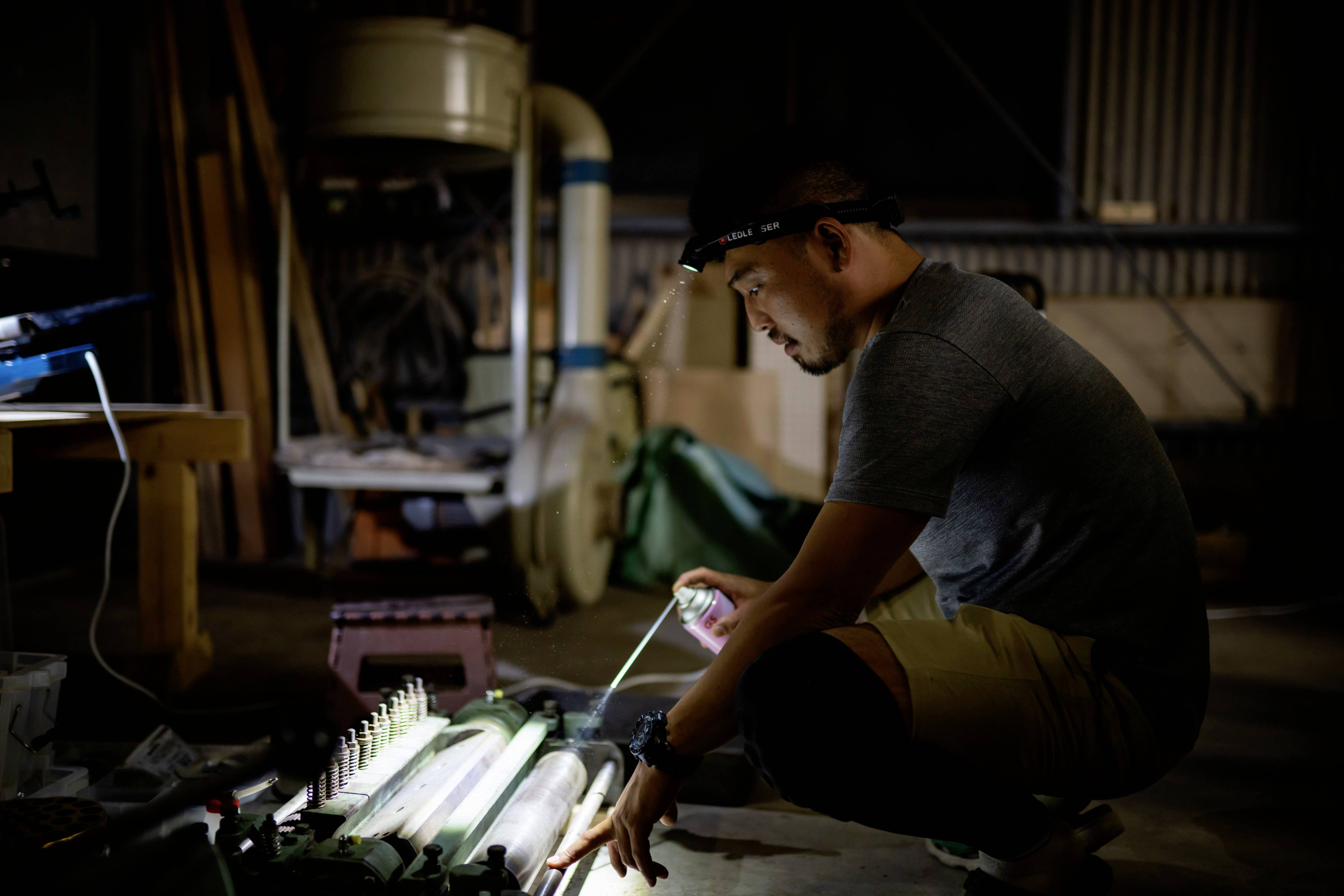 Eine Person arbeitet konzentriert in einer Werkstatt bei schwacher Beleuchtung, untersucht ein Objekt auf dem Boden mit einer Taschenlampe.