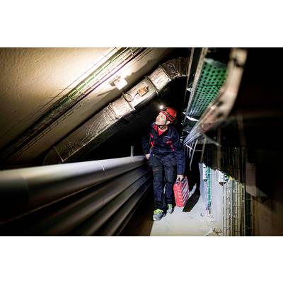 Ein Elektriker mit Helm und Werkzeugkoffer inspiziert Kabel in einem engen Tunnel, beleuchtet von einer Kopflampe.