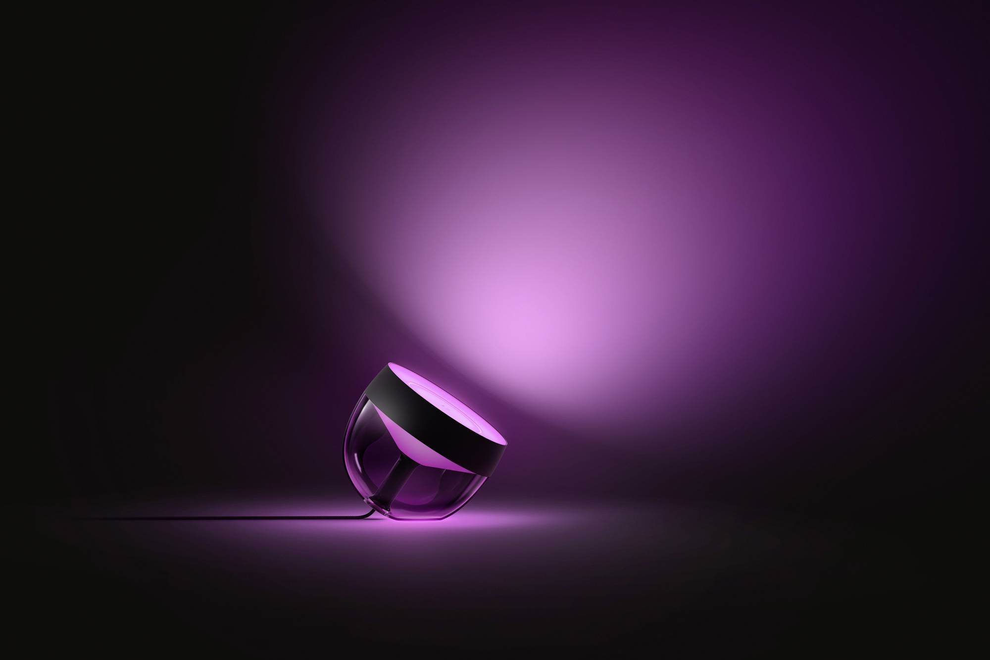 Eine moderne Tischlampe auf der Seite liegend, leuchtet violettes Licht hinter sich auf eine schwarze Fläche, schafft atmosphärische Beleuchtung.