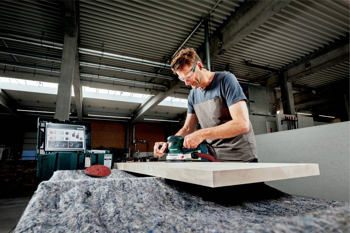 Ein Mann benutzt in einer Werkstatt eine elektrische Schleifmaschine, um ein rechteckiges Holzstück auf einem Tisch zu bearbeiten.