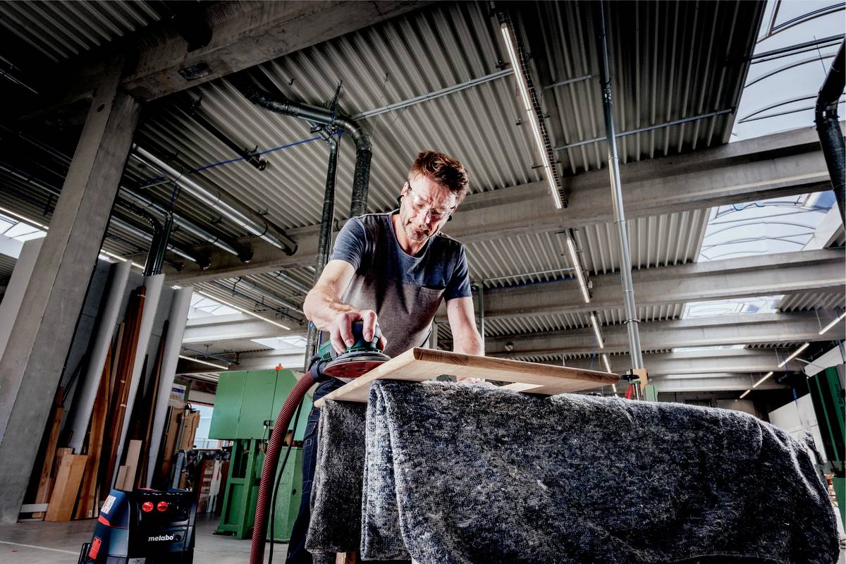 Ein Mann schleift in einer Werkstatt ein Stück Holz auf einem Tisch. Im Hintergrund sind Maschinen und Werkzeuge sichtbar.