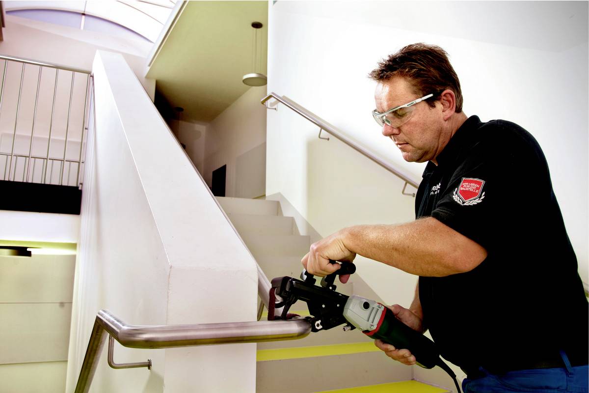 Ein Mann schleift ein Metallgeländer mit einem elektrischen Werkzeug in einem Treppenhaus. Er trägt Schutzbrille und Arbeitskleidung.