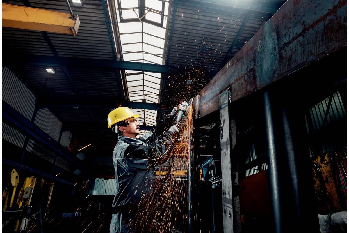 Arbeiter mit Schutzhelm schneidet Metall mit Funkenflug in einer Fabrikhalle. Industrielle Umgebung mit Hochdach und Stahlträgern.