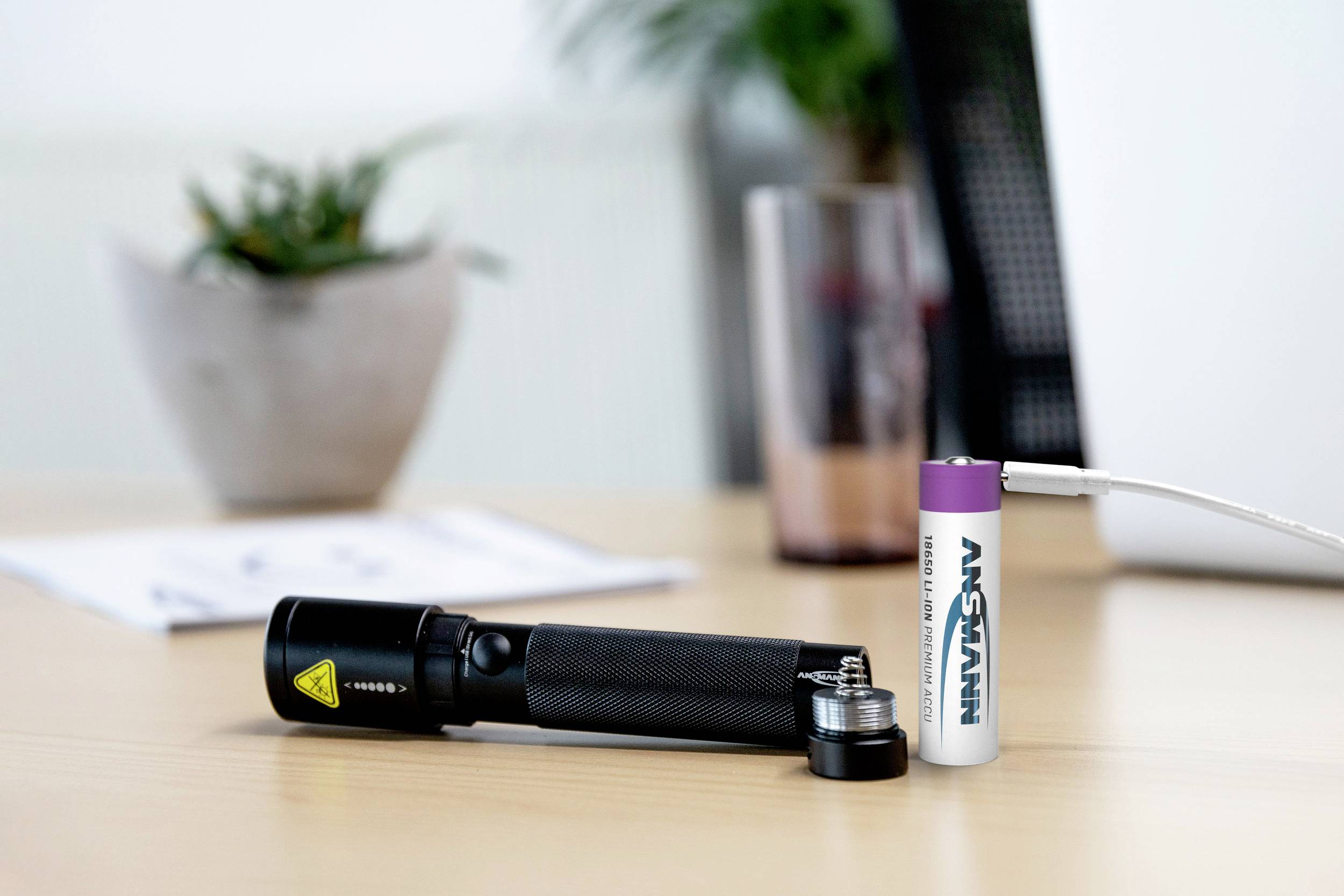 Eine schwarze Taschenlampe liegt auf einem Tisch, daneben eine abgelegte Batterie und ein USB-Kabel. Im Hintergrund ist eine Pflanze zu sehen.