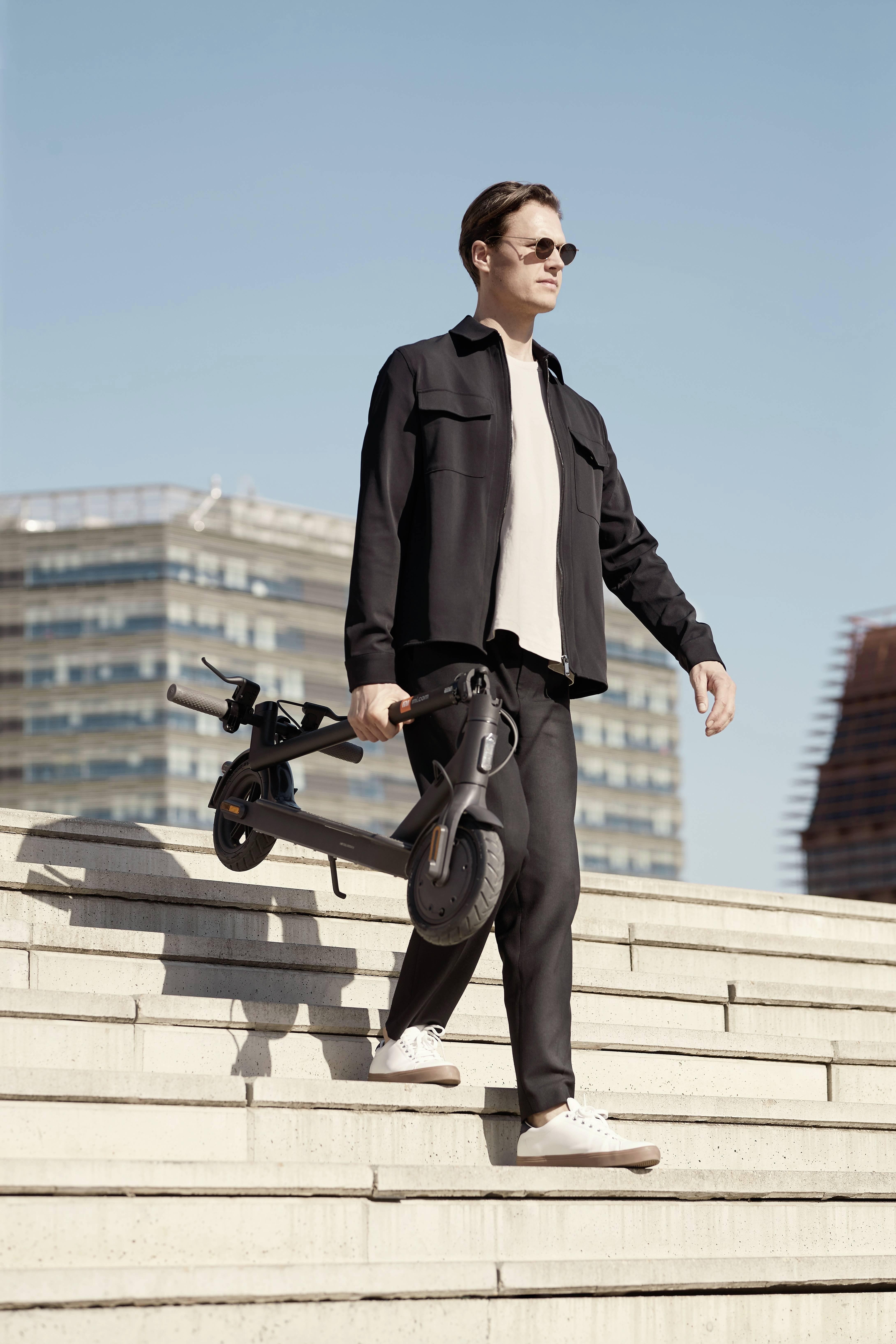 Ein Mann in schwarzer Kleidung geht eine Treppe hinunter und trägt einen E-Scooter. Im Hintergrund sind moderne Gebäude zu sehen.