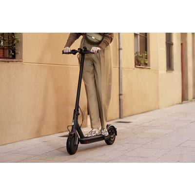 Person fährt auf einem schwarzen Elektroroller eine gepflasterte Straße entlang; Stadtumgebung mit beige Gebäude im Hintergrund.