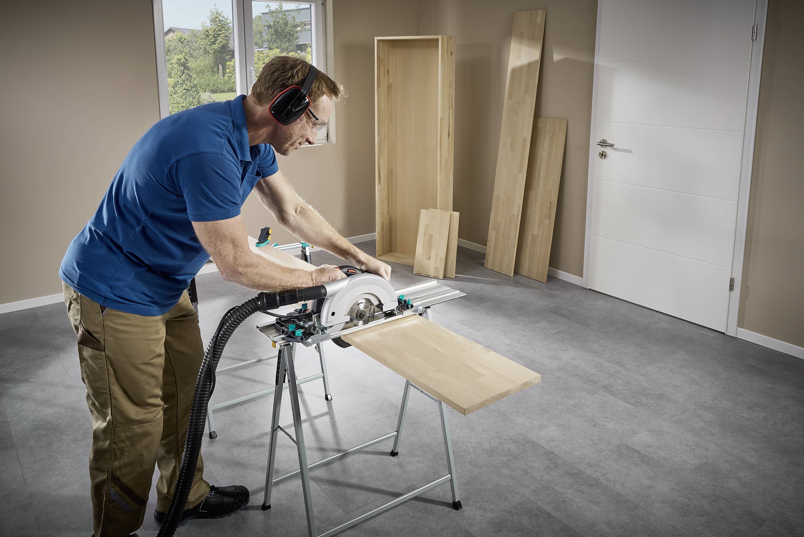 Ein Mann in Schutzbrille und Ohrenschützern schneidet ein Holzbrett mit einer Tischkreissäge in einem Zimmer mit Holzmöbeln.