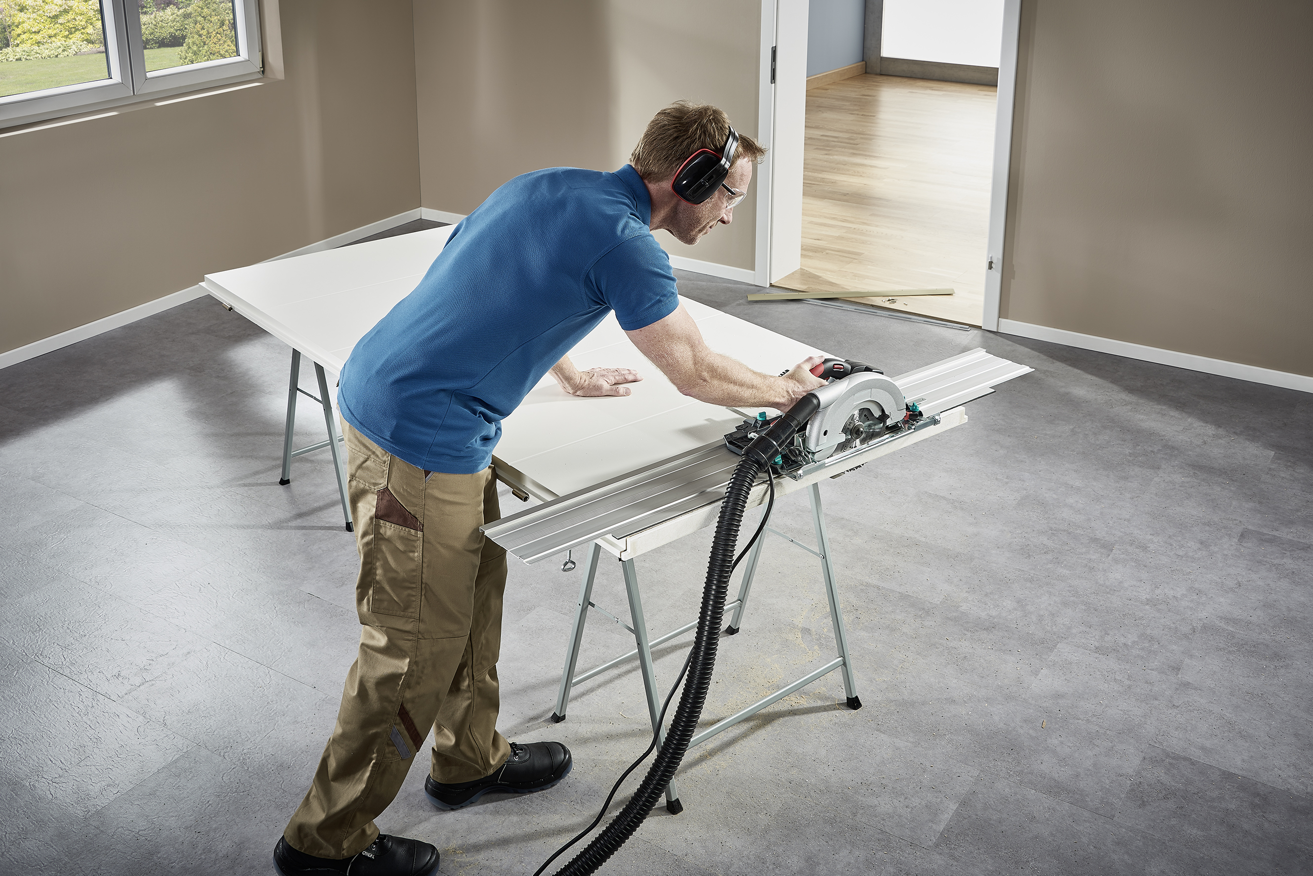 Ein Mann sägt mit einer Kreissäge an einem Tisch in einem leeren Raum, trägt Gehörschutz und Arbeitskleidung.