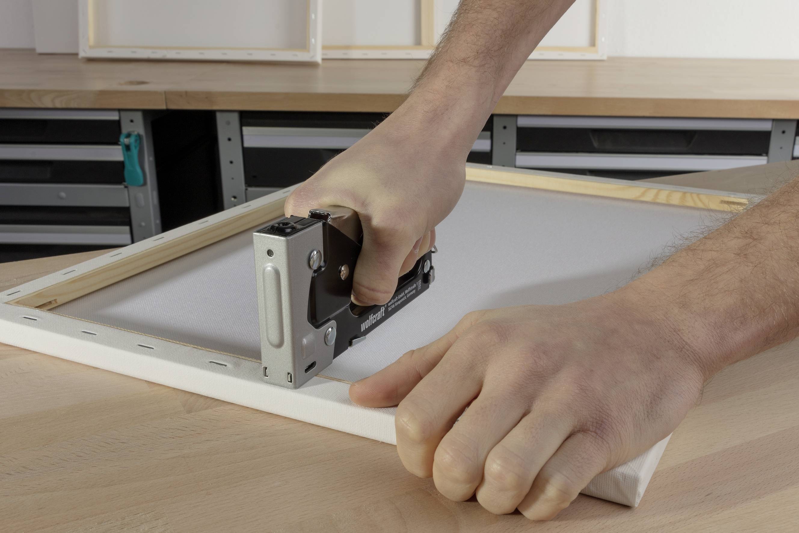 'Zwei Hände befestigen eine Leinwand mit einem Tacker auf einem Holzrahmen. Der Tisch im Hintergrund ist aus Holz.'
