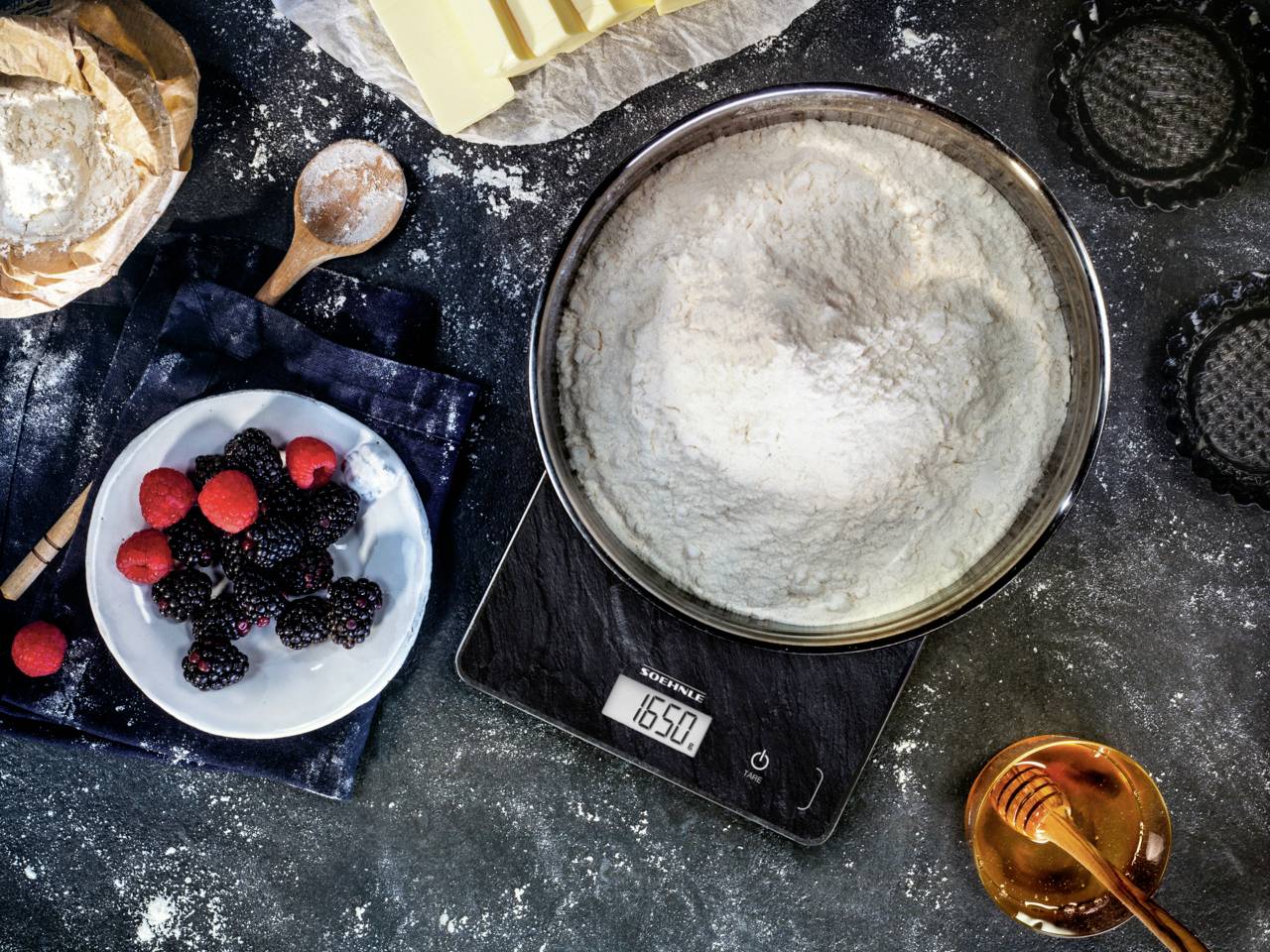 Mehl in einer Schüssel auf einer Waage zeigt 1650 Gramm, umgeben von Beeren, Backformen, einem Holzlöffel, Butter und einer Honigschale.