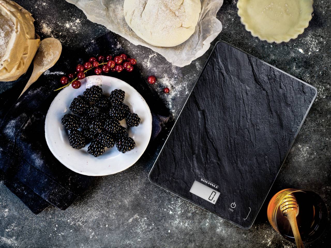 Eine digitale Küchenwaage zeigt '0' an, umgeben von einem Teller mit Brombeeren, roten Beeren, Teig, Honig und einer Holzkelle.