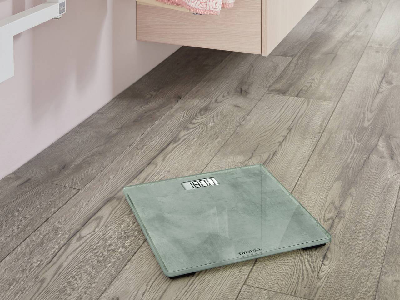 Digitale Personenwaage auf einem Holzboden, zeigt 180.4 kg an. Im Hintergrund sind Möbel erkennbar.