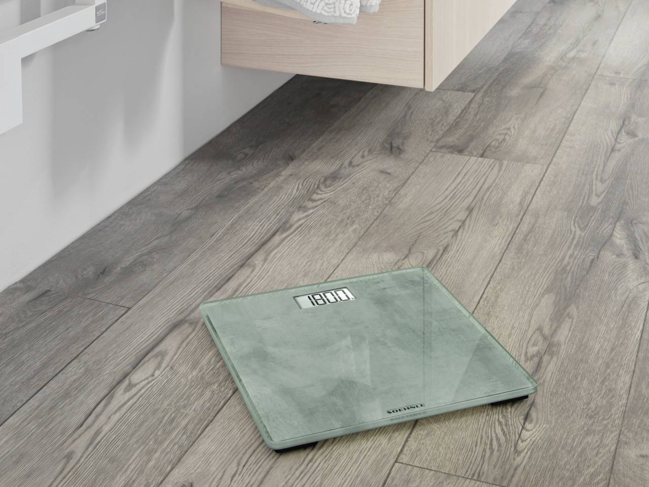 Eine digitale Waage steht auf einem Holzfußboden im Badezimmer, unweit eines hängenden Schranks. Sie zeigt das Gewicht an.