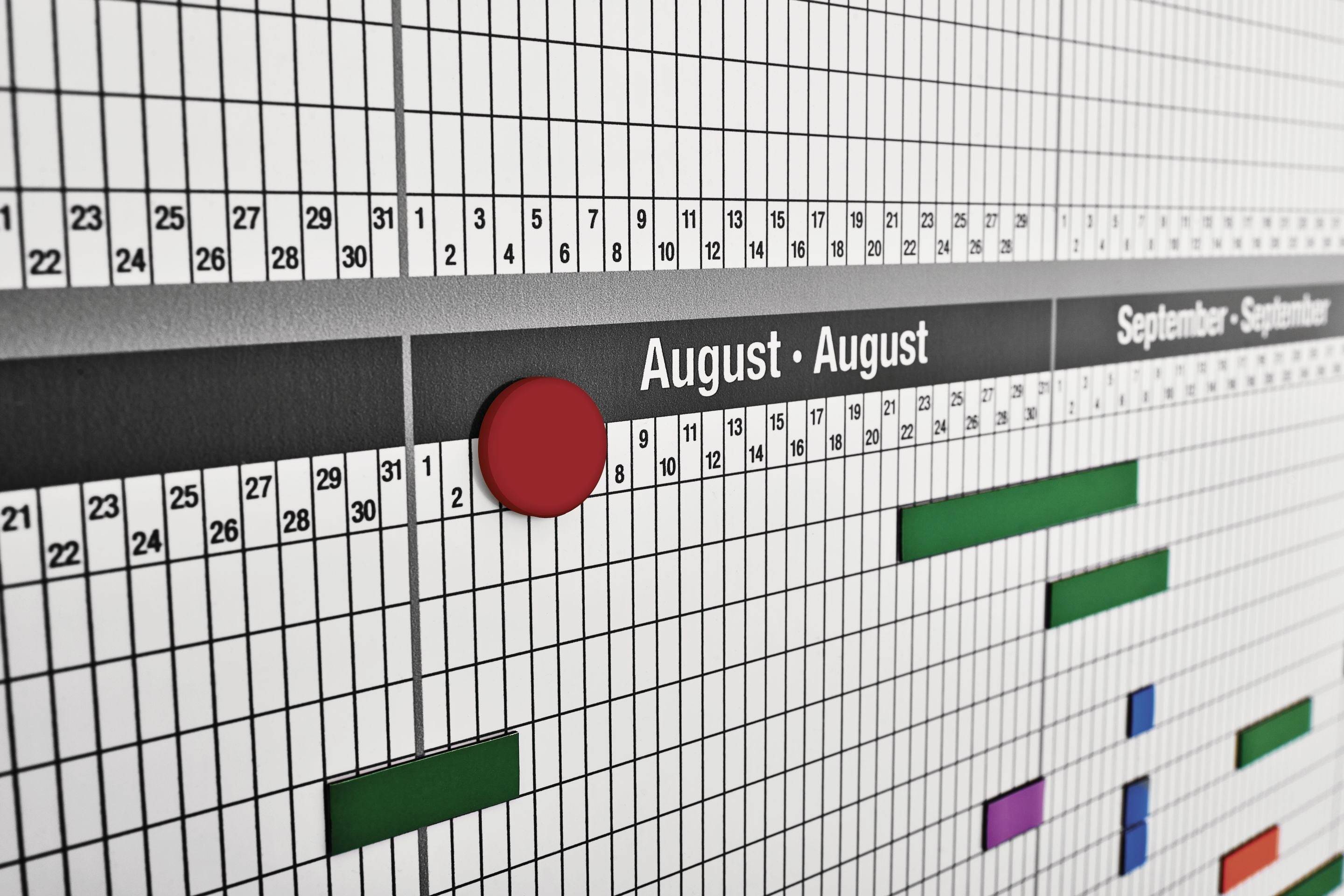 'Wandkalender mit einem roten Markierungspunkt im August und farbigen Balken, die verschiedene Aktivitäten und ihre Zeiträume darstellen.'