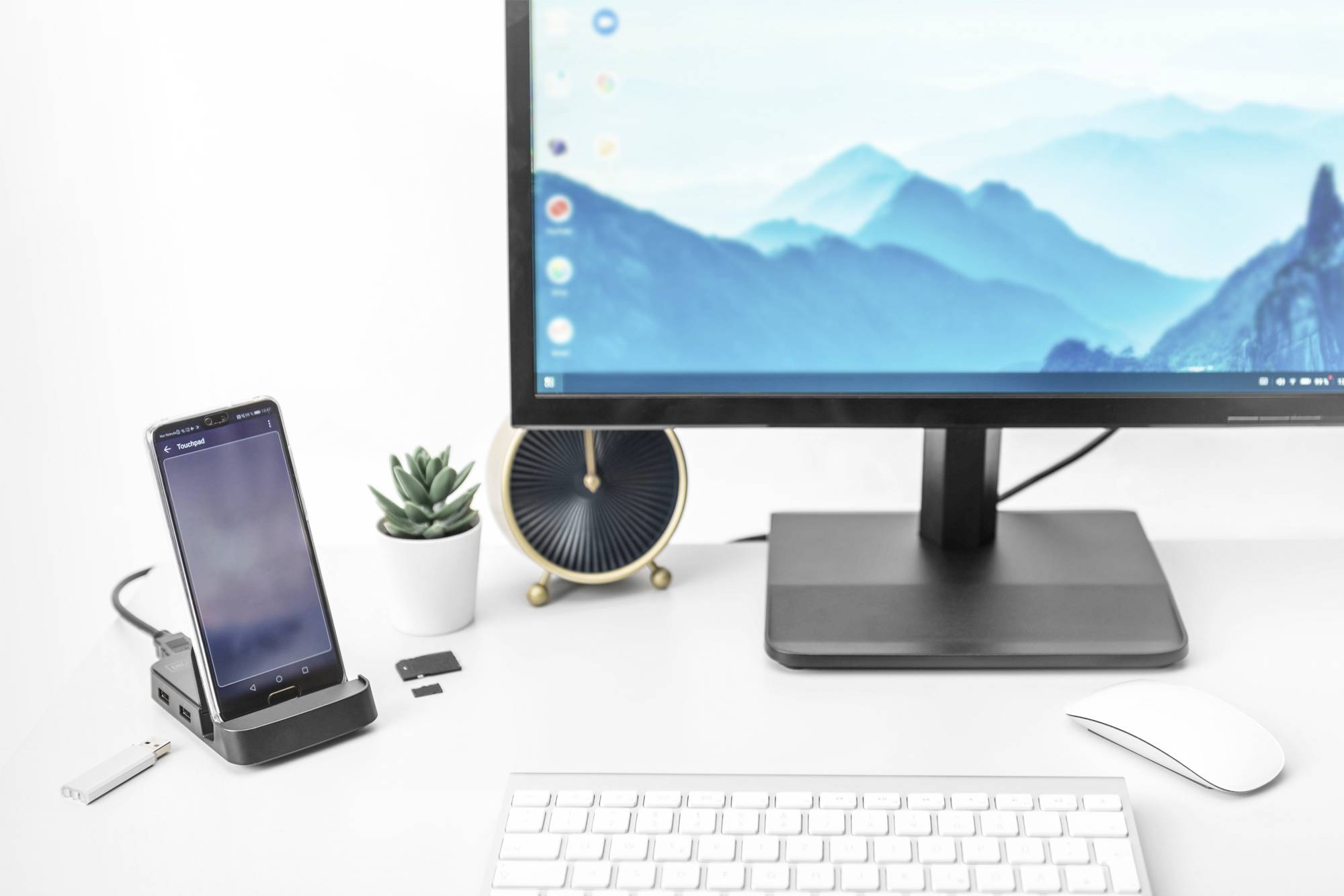 Ein Schreibtisch mit einem Monitor, Smartphone auf einer Dockingstation, USB-Stick, SD-Karten, Tastatur, Maus, Pflanze und Uhr.