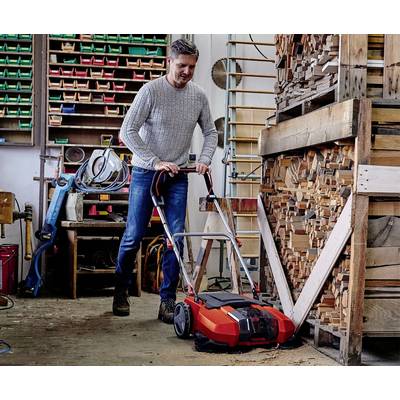 Ein Mann benutzt in einer Werkstatt einen Kehrmaschine, um den Boden in der Nähe eines Holzstapels zu reinigen. Werkzeuge im Hintergrund.