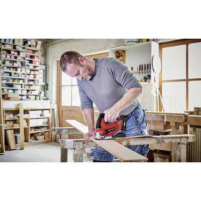 Ein Mann in einer Werkstatt schneidet mit einer roten Stichsäge ein Holzbrett. Im Hintergrund sind Regale mit Werkzeug und Materialien.