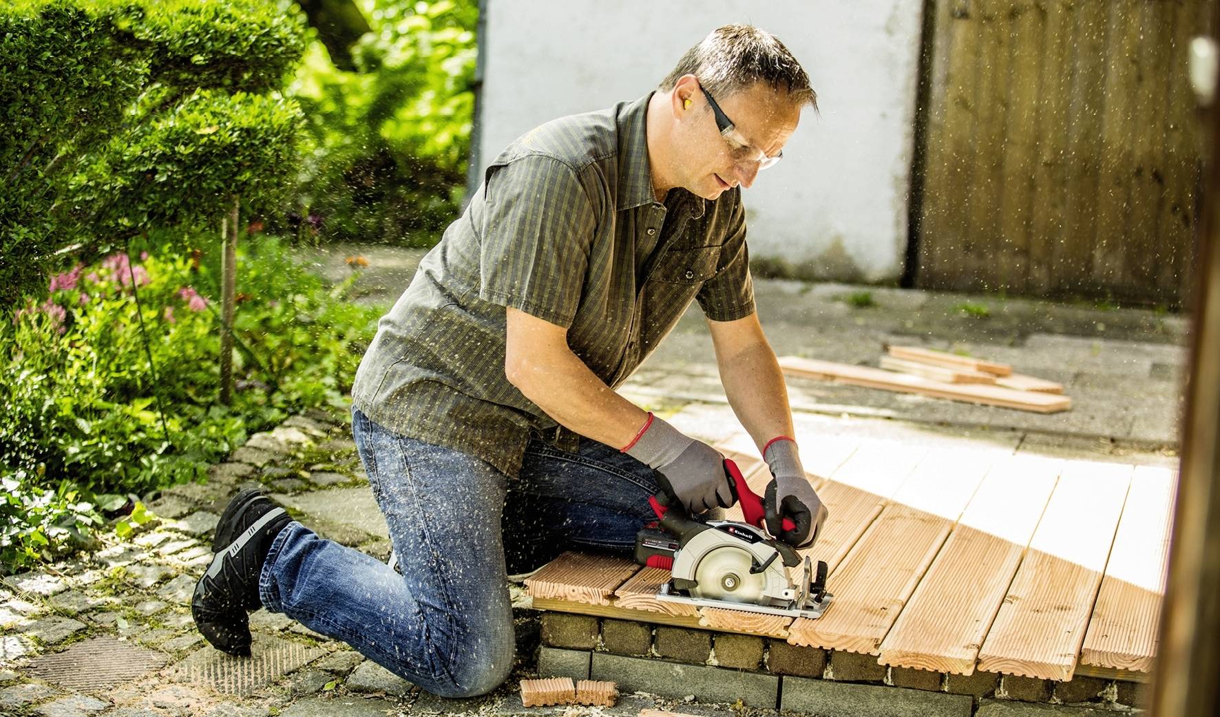 Ein Mann kniet im Freien und schneidet mit einer Kreissäge Holzdielen. Er trägt Schutzbrille und Handschuhe. Umgebung: Garten.
